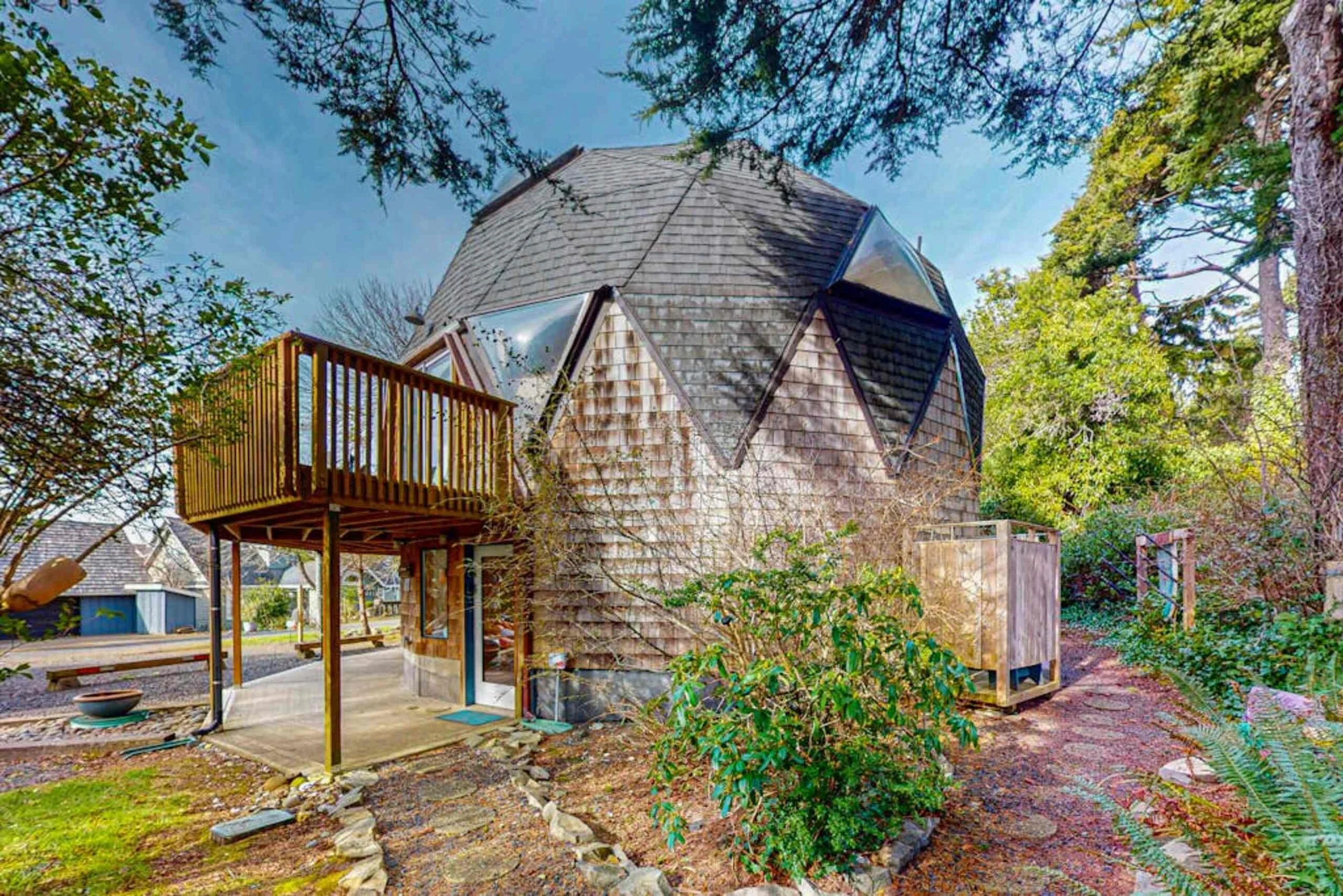 A small house with a rounded wooden exterior and a shingled roof, featuring a balcony and a patio, surrounded by trees and outdoor space.