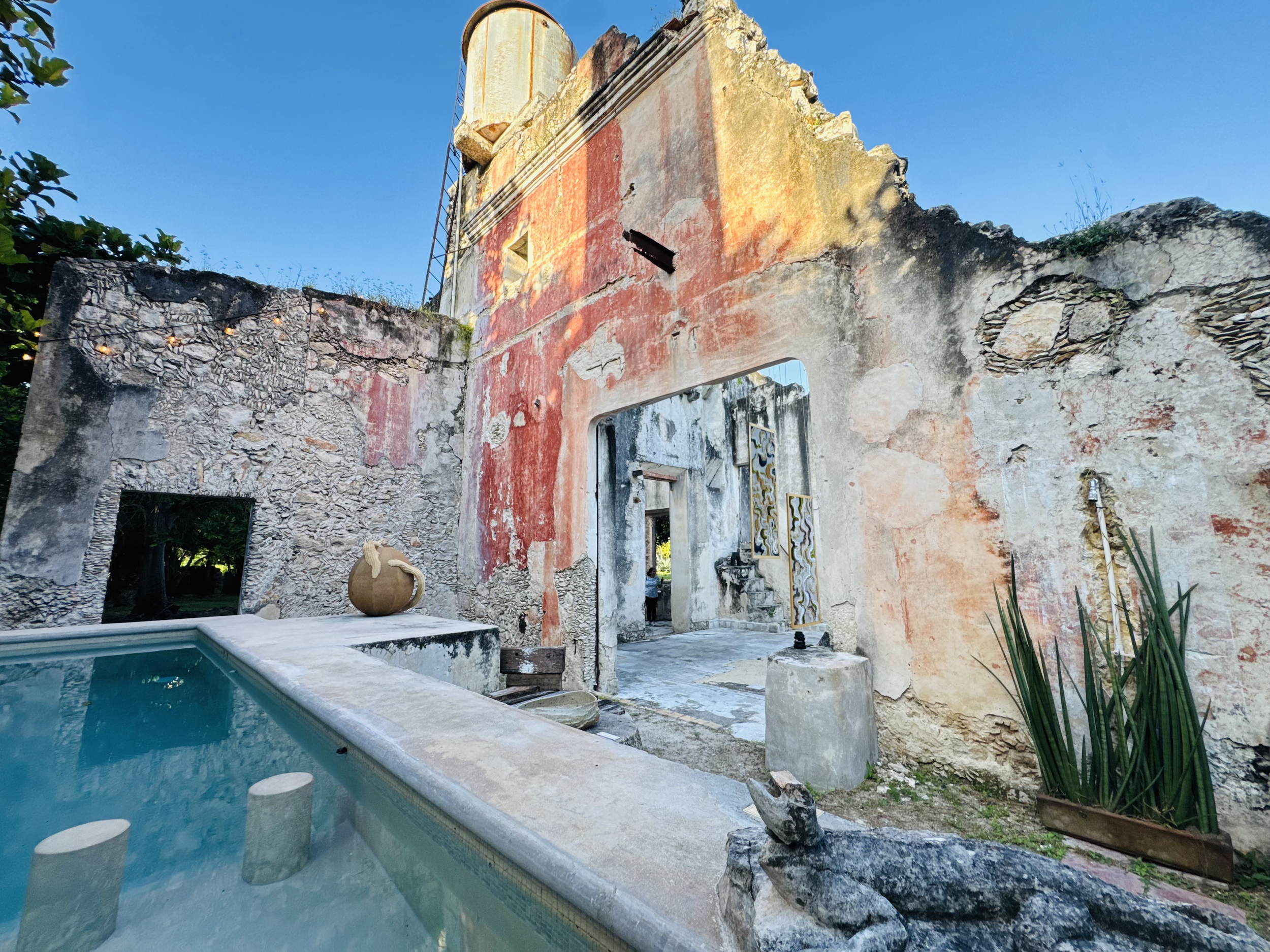 Ruined old building with a weathered, faded painted wall, an archway, and a dripstone on the side, next to a swimming pool with water and steps leading into it.