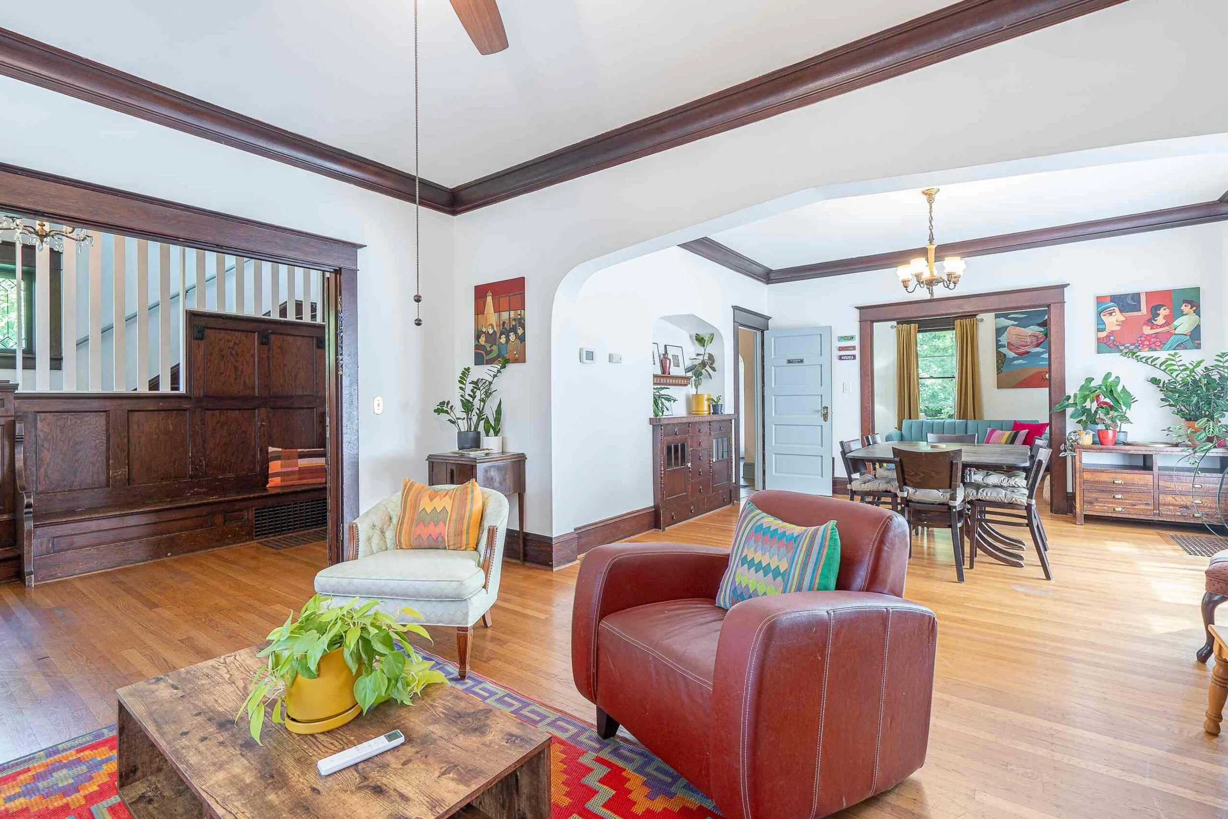 Living room with colorful furniture, wooden floors, wall art, plants, and a ceiling fan, with walls painted white and framed by dark wood trim.