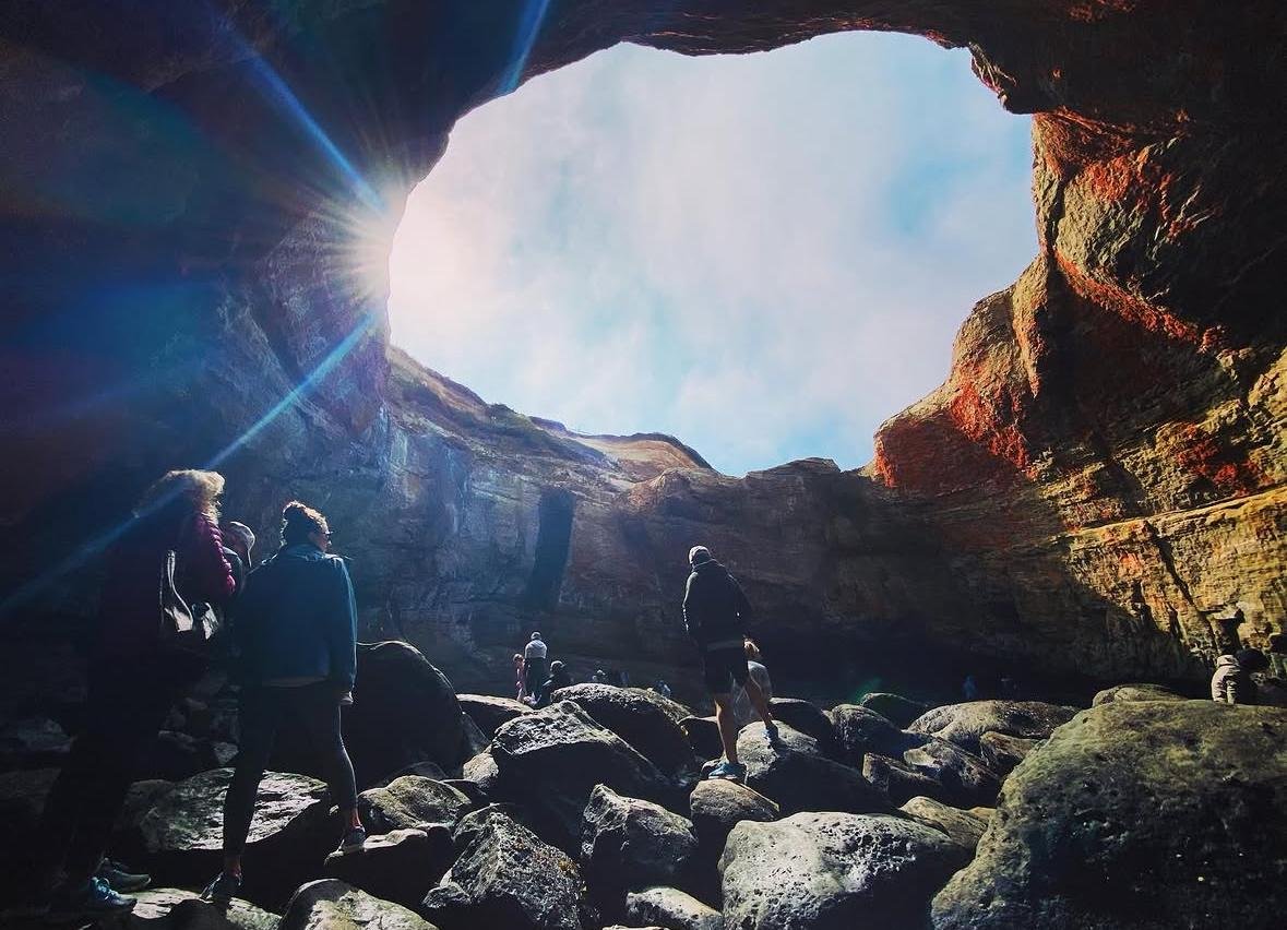 People exploring inside a large cave with a wide opening letting in sunlight, rocky floor, and a blue sky visible through the opening.