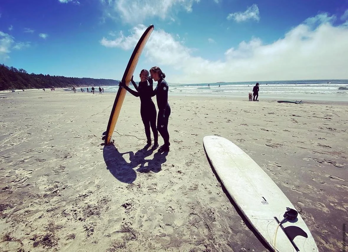 Two women in wetsuits standing on a sandy beach holding a surfboard, with a white surfboard lying on the sand near them and the ocean waves in the background.