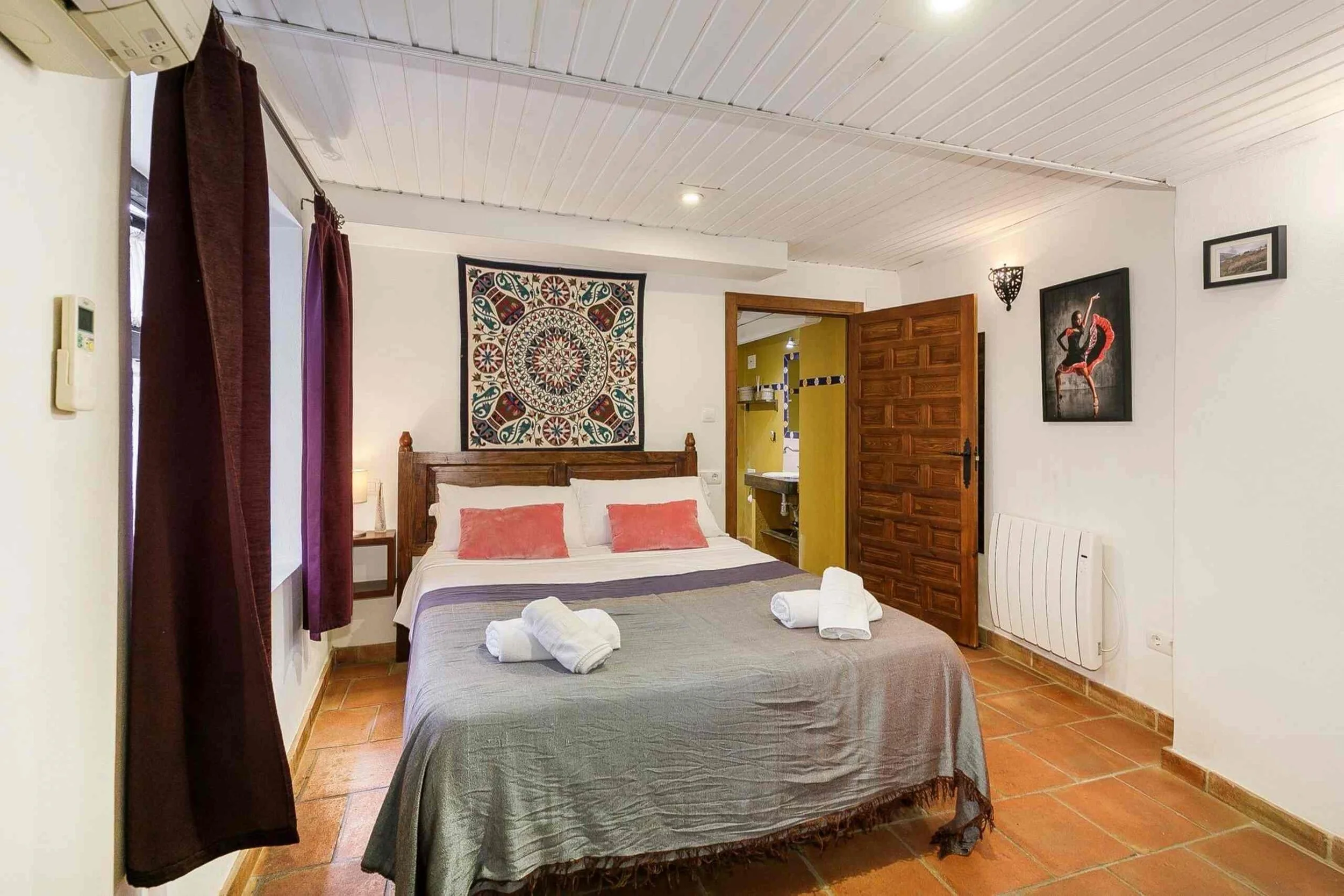 A bedroom with two beds, one with multiple pillows, and other furniture including wall art, curtains, and a bathroom visible through an open door.