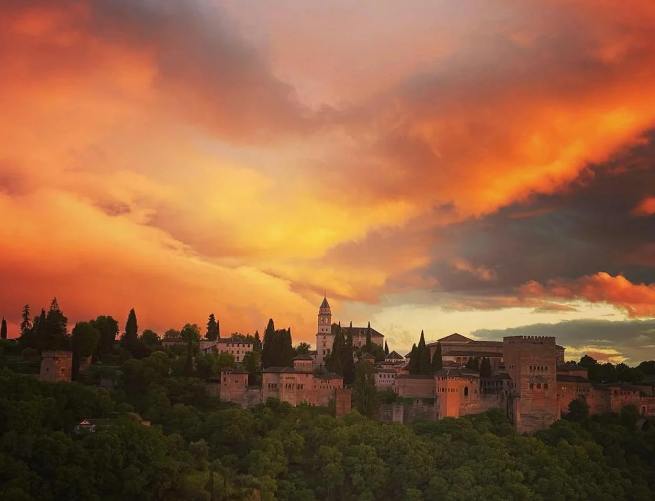 Sunset over a historic hilltop castle with a tall bell tower, surrounded by lush green trees and a dramatic orange and pink sky.