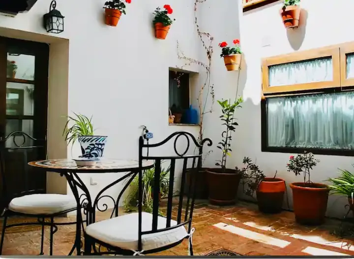 Outdoor patio with black metal table and chairs, potted plants, and white walls decorated with hanging flower pots and vine, sunny day lighting.