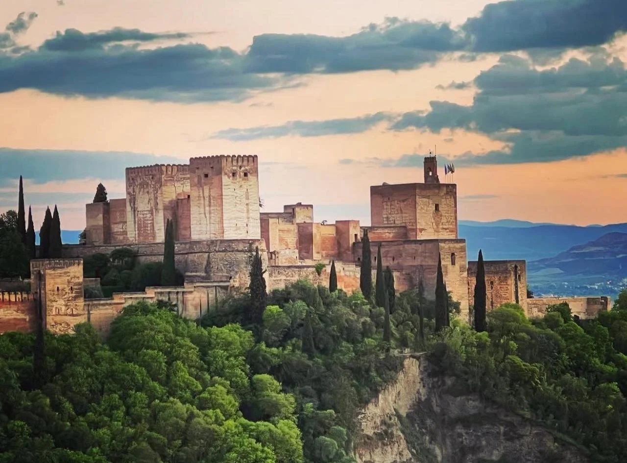 A historic castle with tall stone towers and walls situated on a hill surrounded by lush green trees, under a partly cloudy sky during sunset.