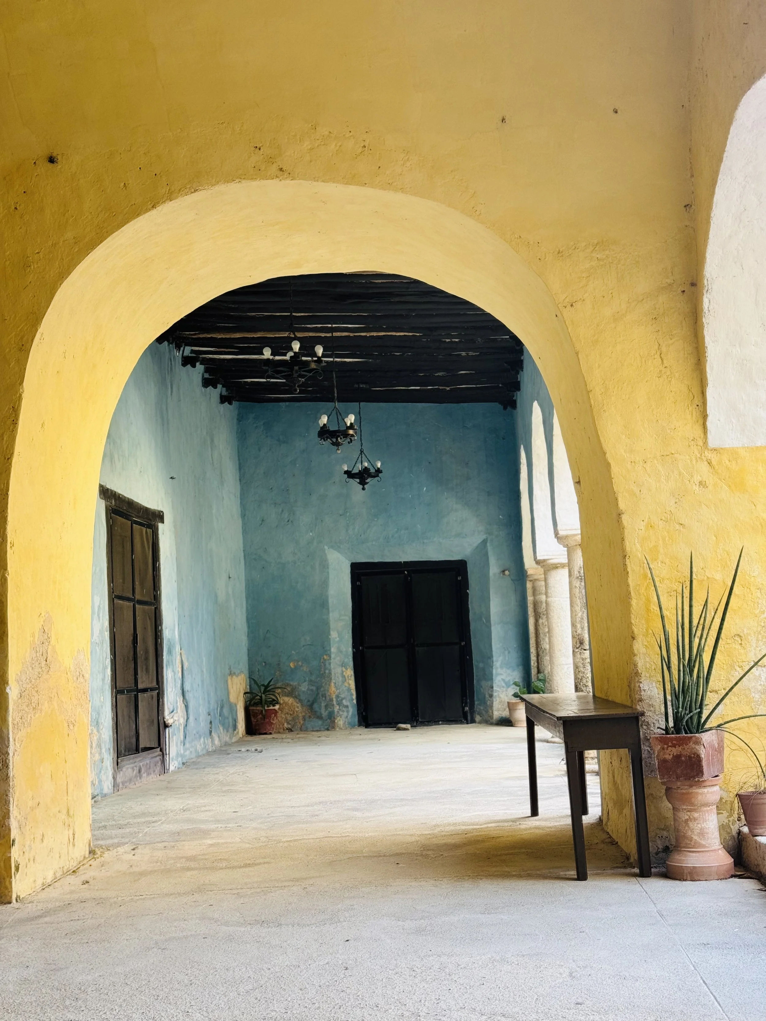 A courtyard with yellow arched walls, a blue wall inside, black doors, hanging chandeliers, and potted plants.