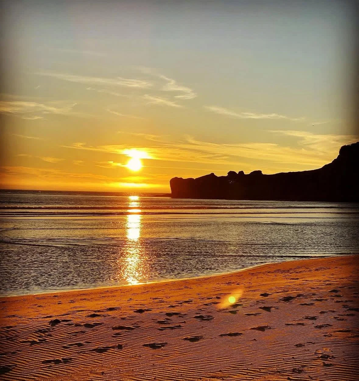 Sunset over the ocean with a rocky coastline in the distance, calm water reflecting the sunlight, sandy beach with footprints in the foreground.