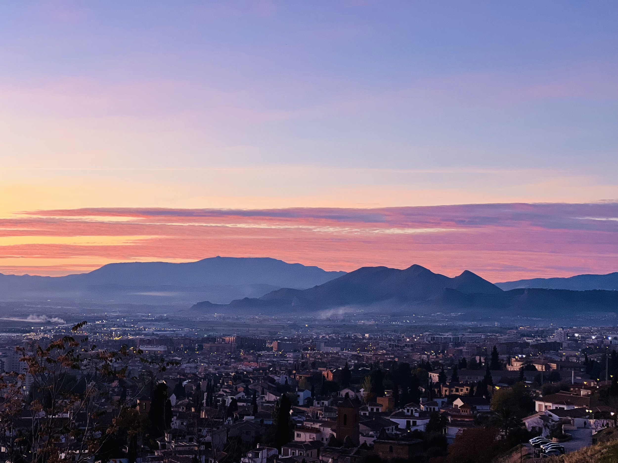 A cityscape at sunset with mountains in the background and a colorful sky in shades of purple, pink, and orange.