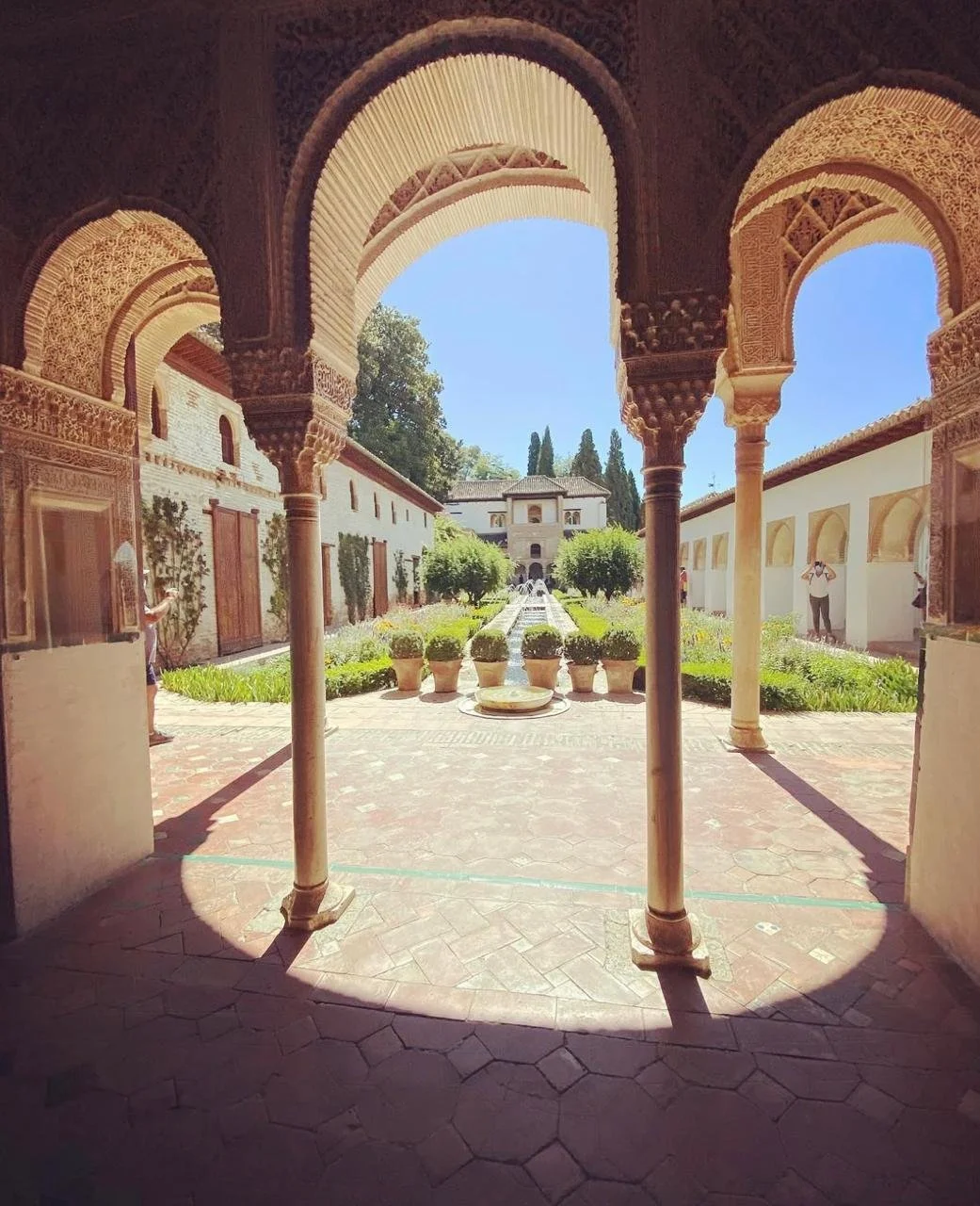 View of a courtyard garden with potted plants, fountains, and trees, seen through decorative arches of Islamic architecture.