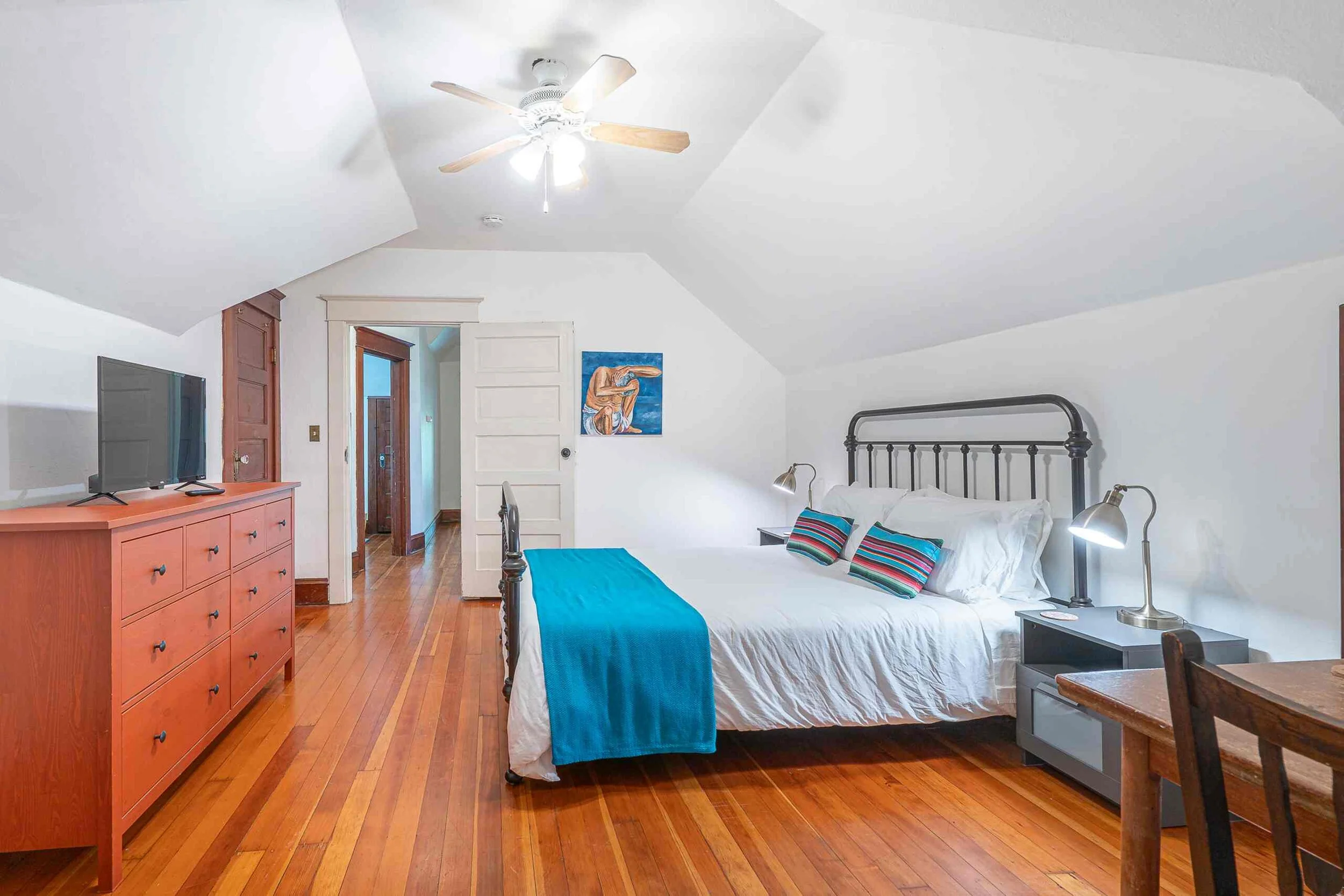 A cozy bedroom with white walls and a vaulted ceiling, featuring a black metal bed with white bedding and colorful striped pillows, a wooden dresser with a flat-screen TV, a small nightstand with a silver desk lamp, and a painting of an Indigenous pe