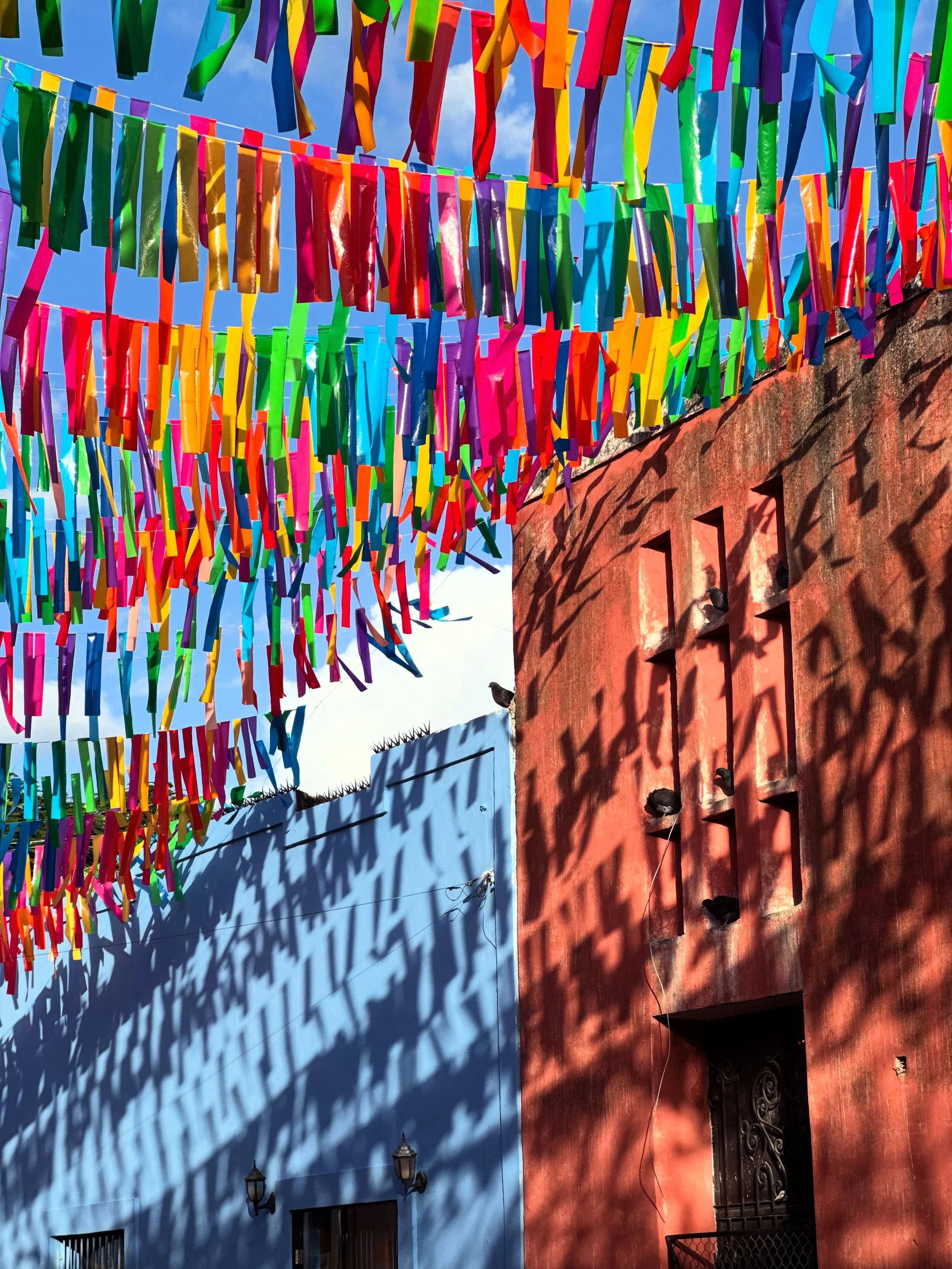 Colorful paper streamers hanging across an outdoor space under a blue sky, casting shadows on a red wall.