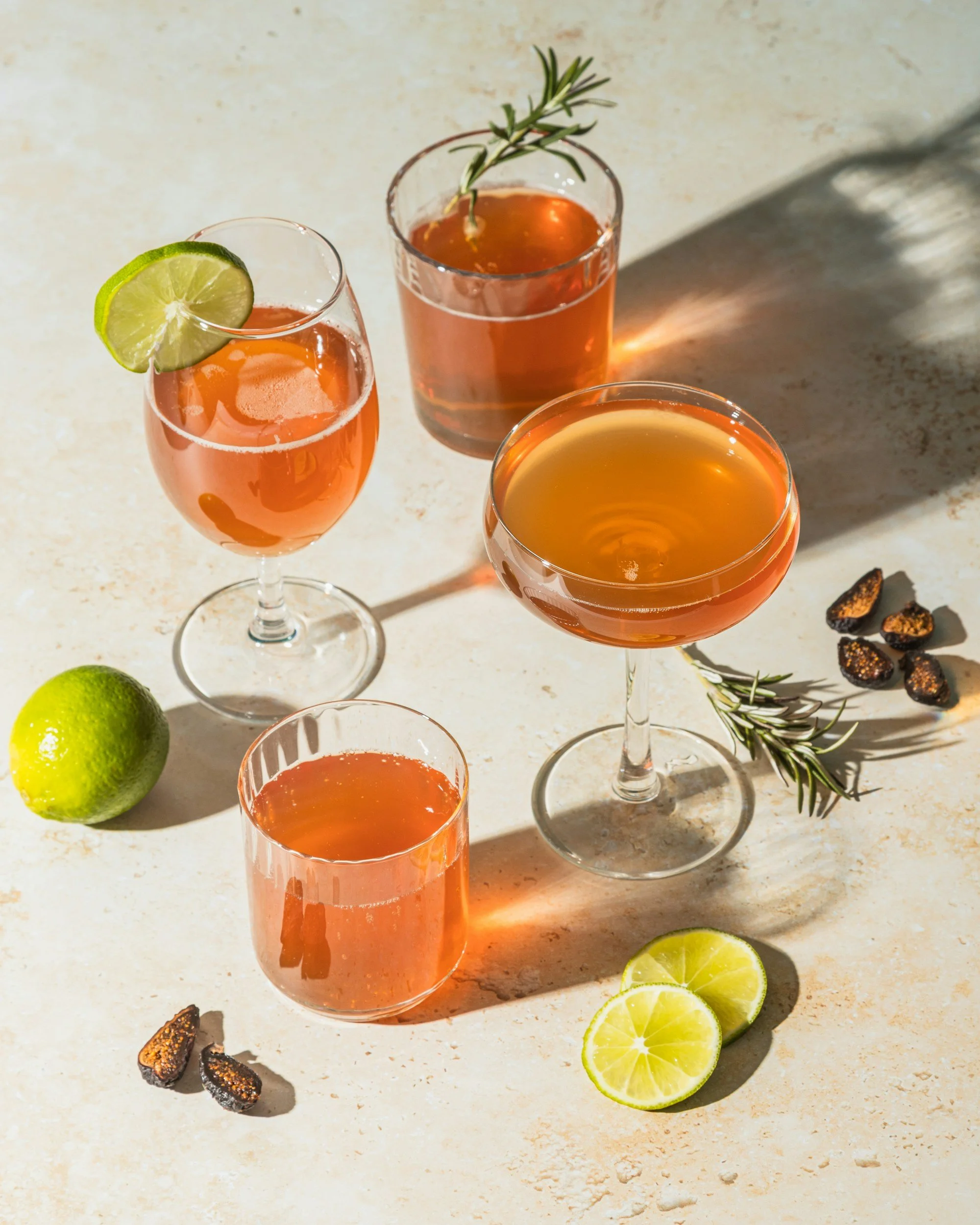 Three cocktail glasses with pinkish-orange drinks on a light-colored surface, garnished with lime slices and rosemary sprigs. There are whole and sliced limes, and dried fruit pieces nearby, with shadows cast on the surface.