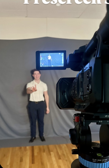 A man standing in front of a gray backdrop, being filmed with a professional camera.