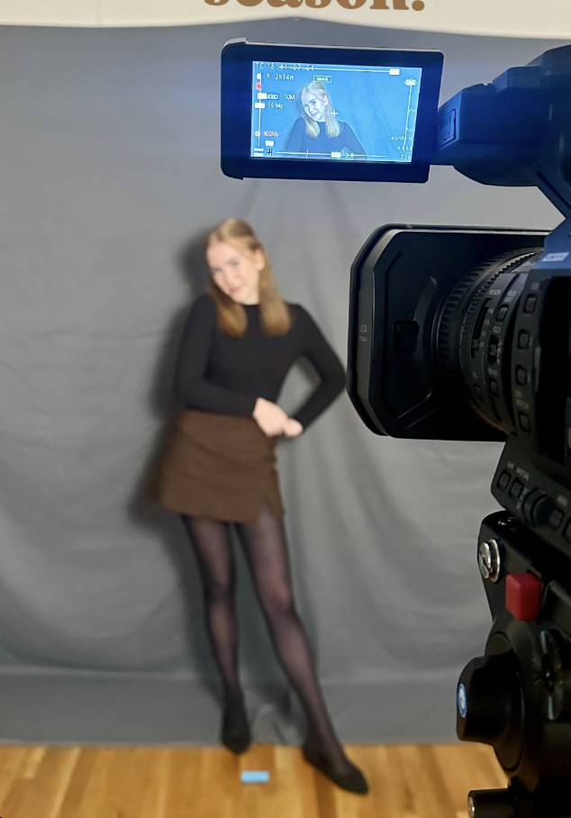 A woman stands in front of a gray backdrop, dressed in a black top, brown skirt, black tights, and shoes, posing with one hand on her hip. A camera on a tripod captures her, with its screen showing her portrait.
