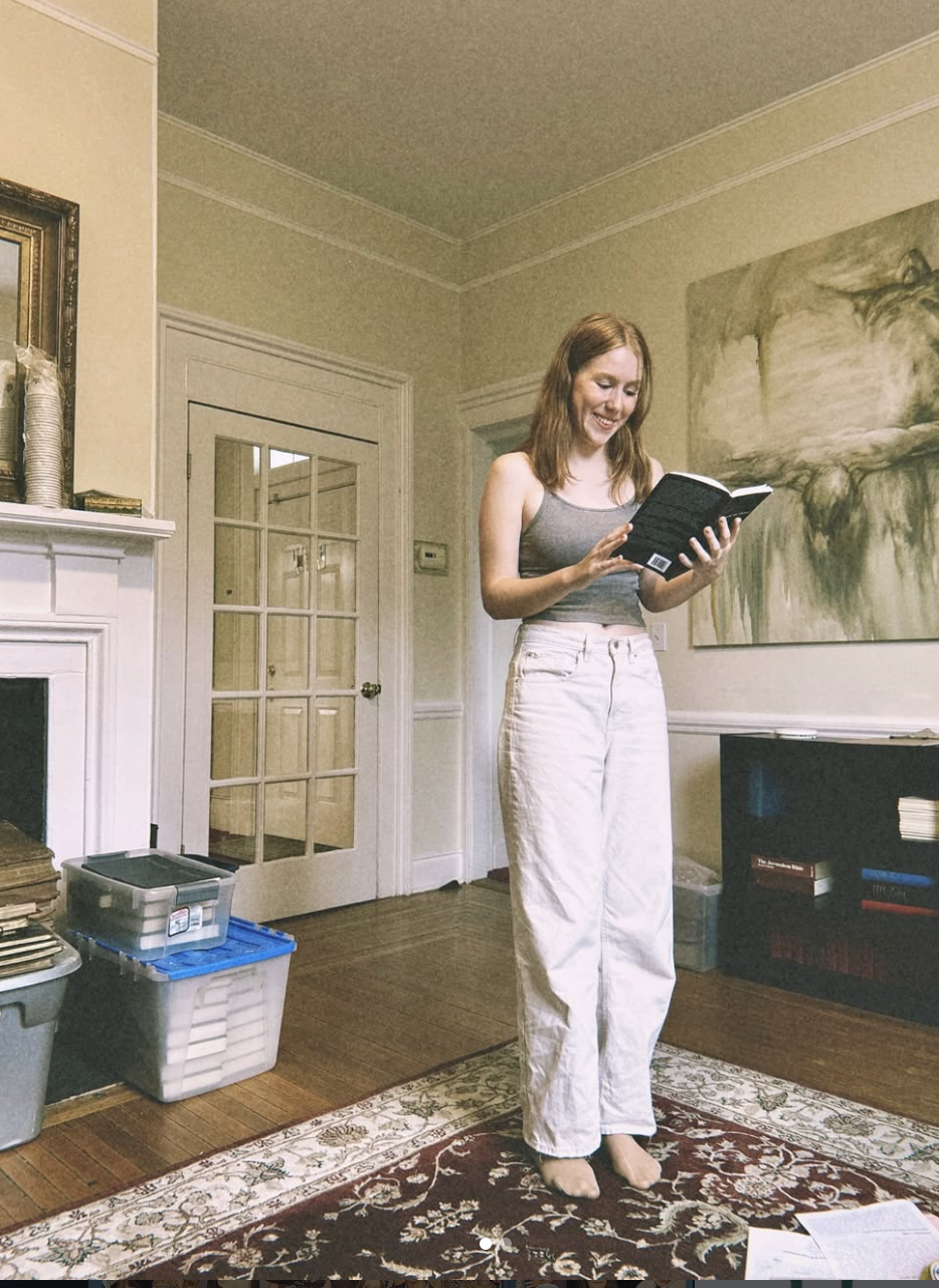 A woman with red hair, wearing a gray tank top and white pants, standing barefoot on a patterned rug in a rehearsal room, reading a play and smiling.