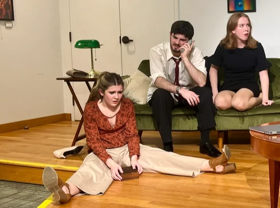 Three young adults sitting on the floor and a couch in a living room. The girl in the foreground is sitting with her legs stretched out, looking distressed. The man in the middle is sitting on the couch, talking on the phone, with a serious expression. The girl on the right is sitting on the couch, looking surprised or shocked. There is a wooden side table with a green lamp and a background with closed white doors.