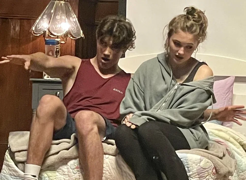 A young man and woman sitting on a bed in a staged bedroom. The man is extending one arm outward and has a shocked expression, while the woman is looking down and slightly to the side, with her hair in a messy bun and wearing casual clothing