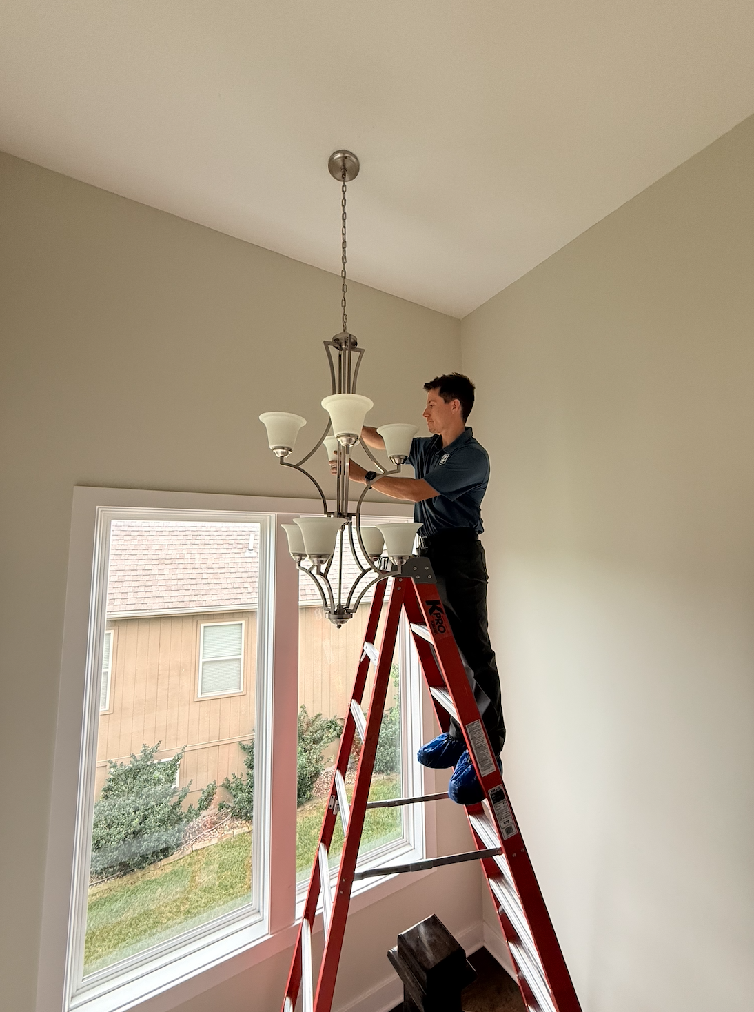 Worker on a red ladder installing a chandelier in a room with large windows and white walls.