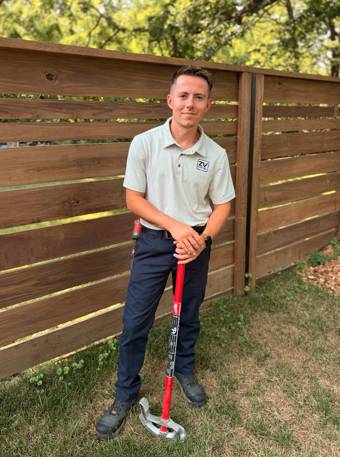 A man standing outdoors in front of a wooden fence, holding a gardening tool or equipment, wearing a light-colored polo shirt, dark pants, and hiking shoes.