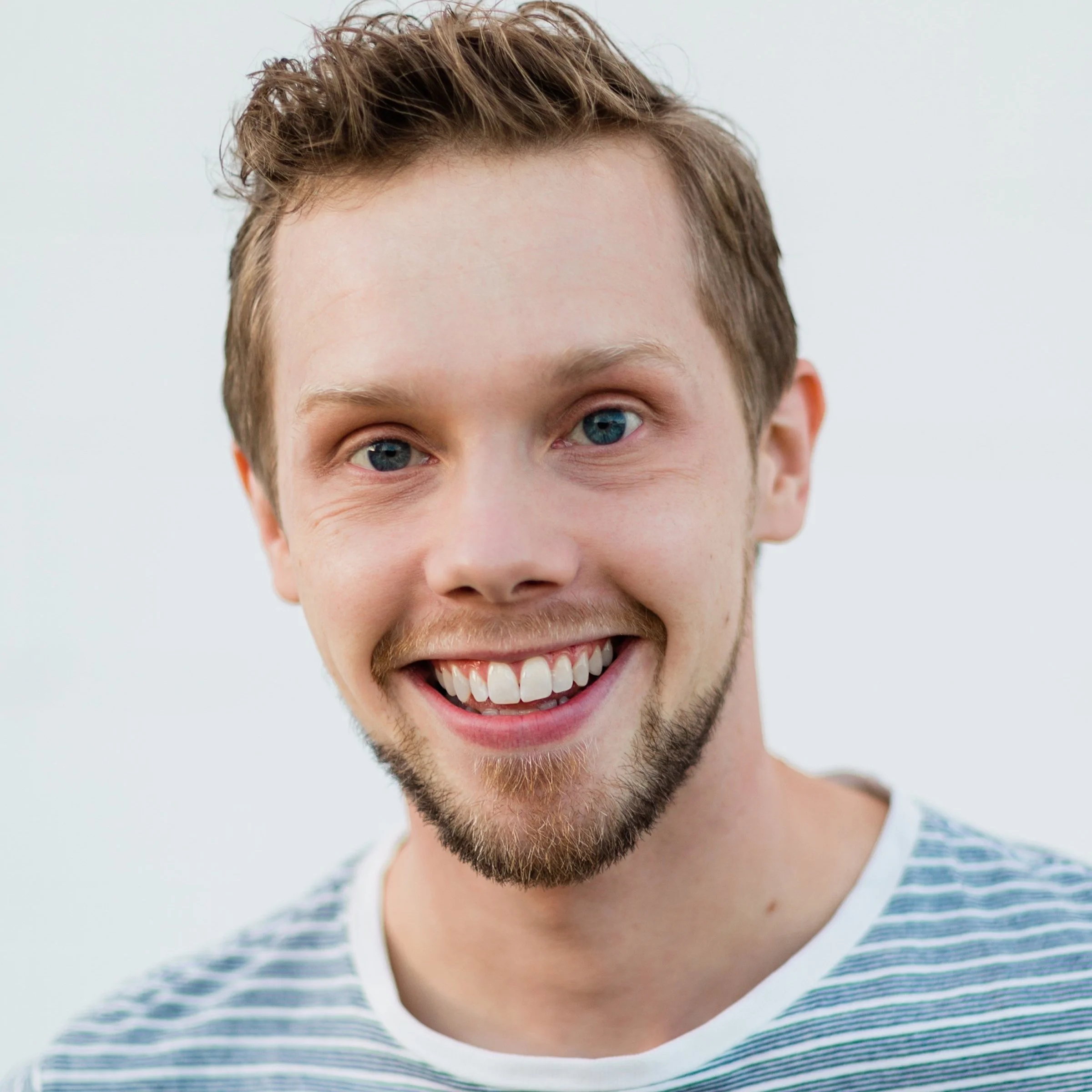 Close-up of a smiling young man with blue eyes, light skin, short wavy brown hair, and light facial hair, wearing a white and gray striped shirt.