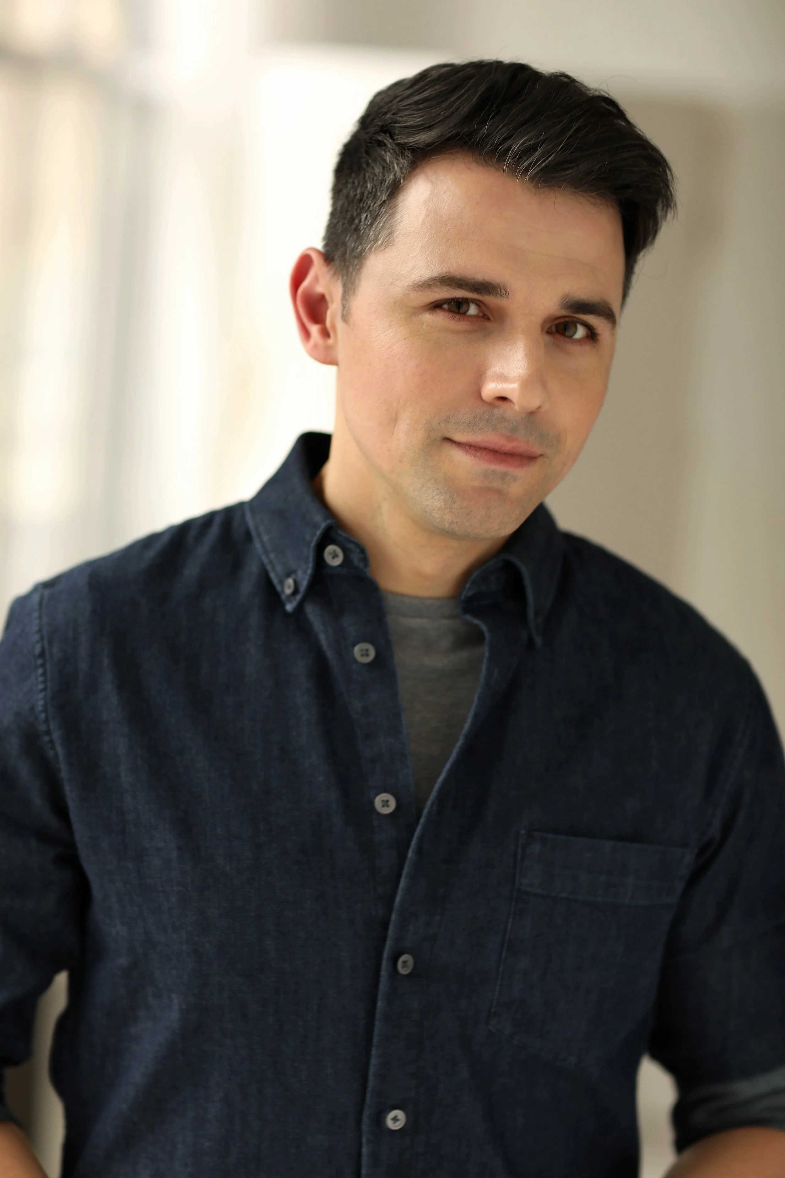 A man with dark brown hair, fair skin, and brown eyes, wearing a dark denim shirt over a gray t-shirt, smiling softly at the camera in a well-lit indoor setting.