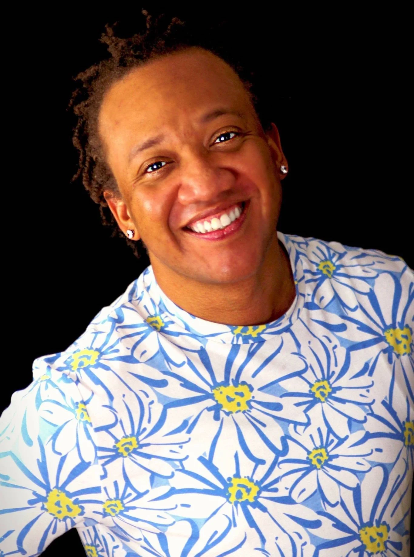 A smiling person with short curly hair, wearing a blue and white floral-patterned shirt, earrings, and standing against a black background.