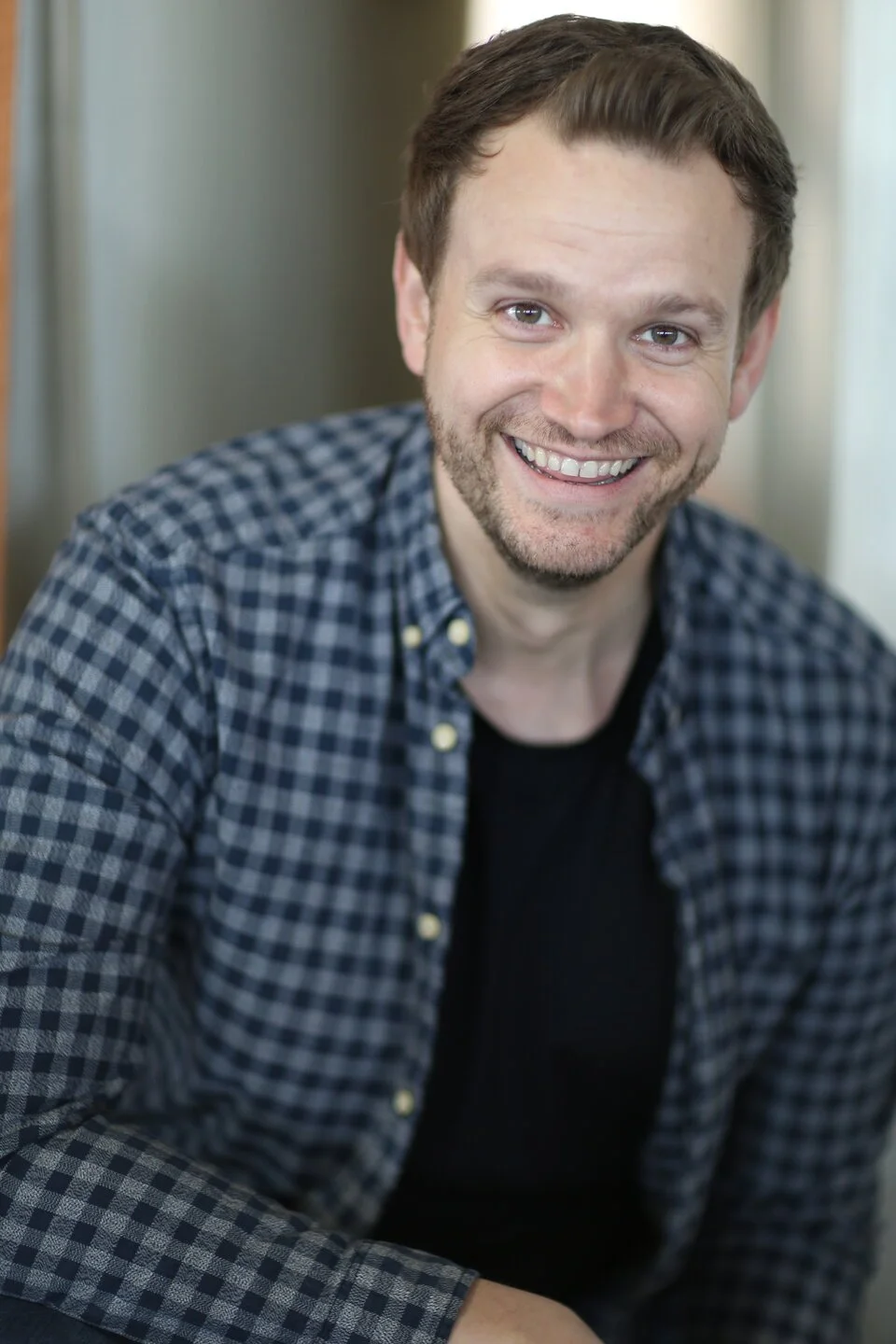 Man with short brown hair and beard smiling, wearing a black shirt and checkered blue and gray shirt, sitting indoors.