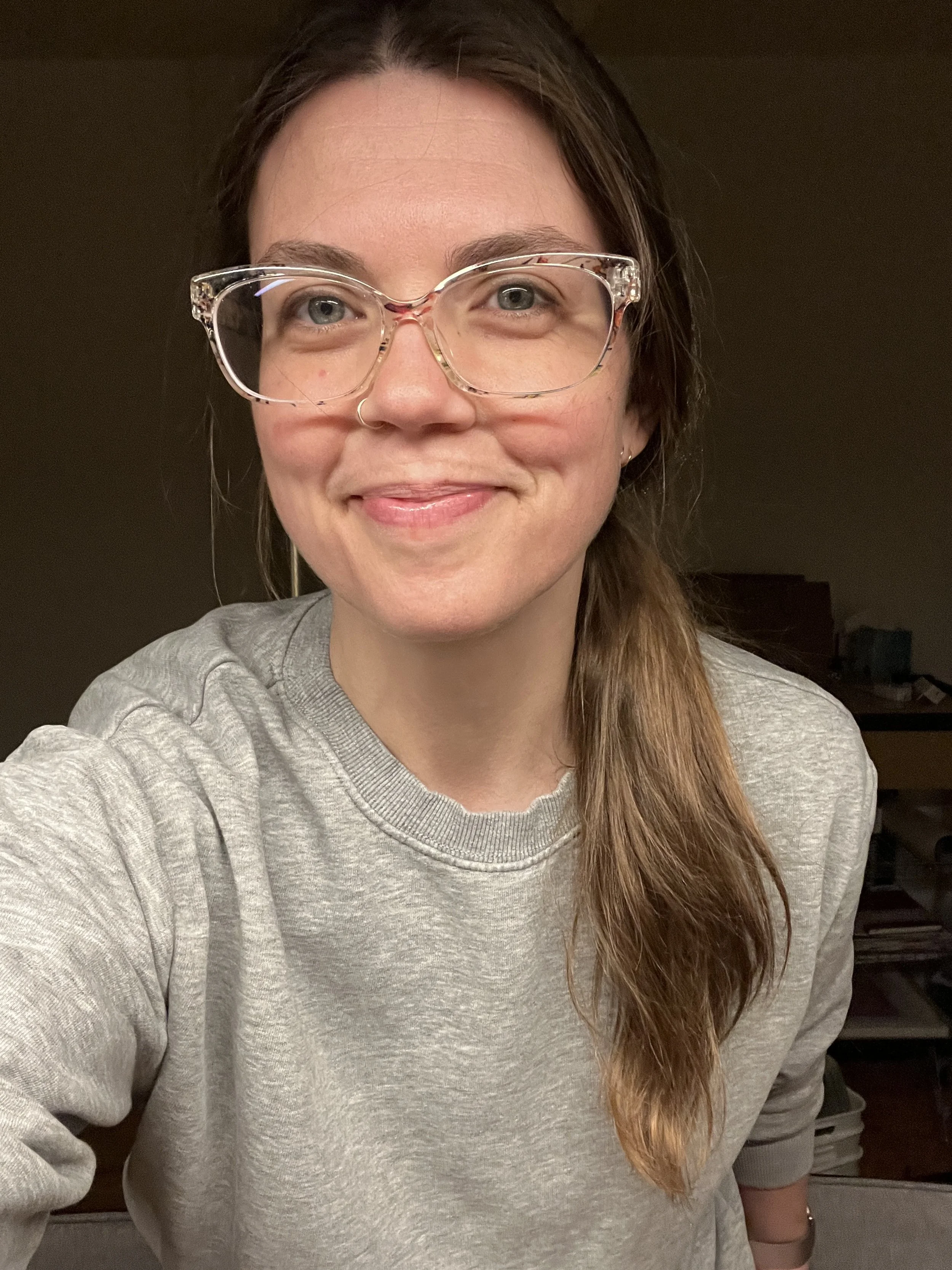 A woman with long, brown hair tied to the side, wearing clear eyeglasses, a nose ring, and a gray sweatshirt, smiling at the camera in a warmly lit room.