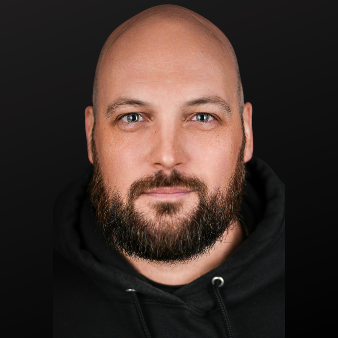 Close-up portrait of a bald man with a beard, wearing a black hoodie, against a dark background.