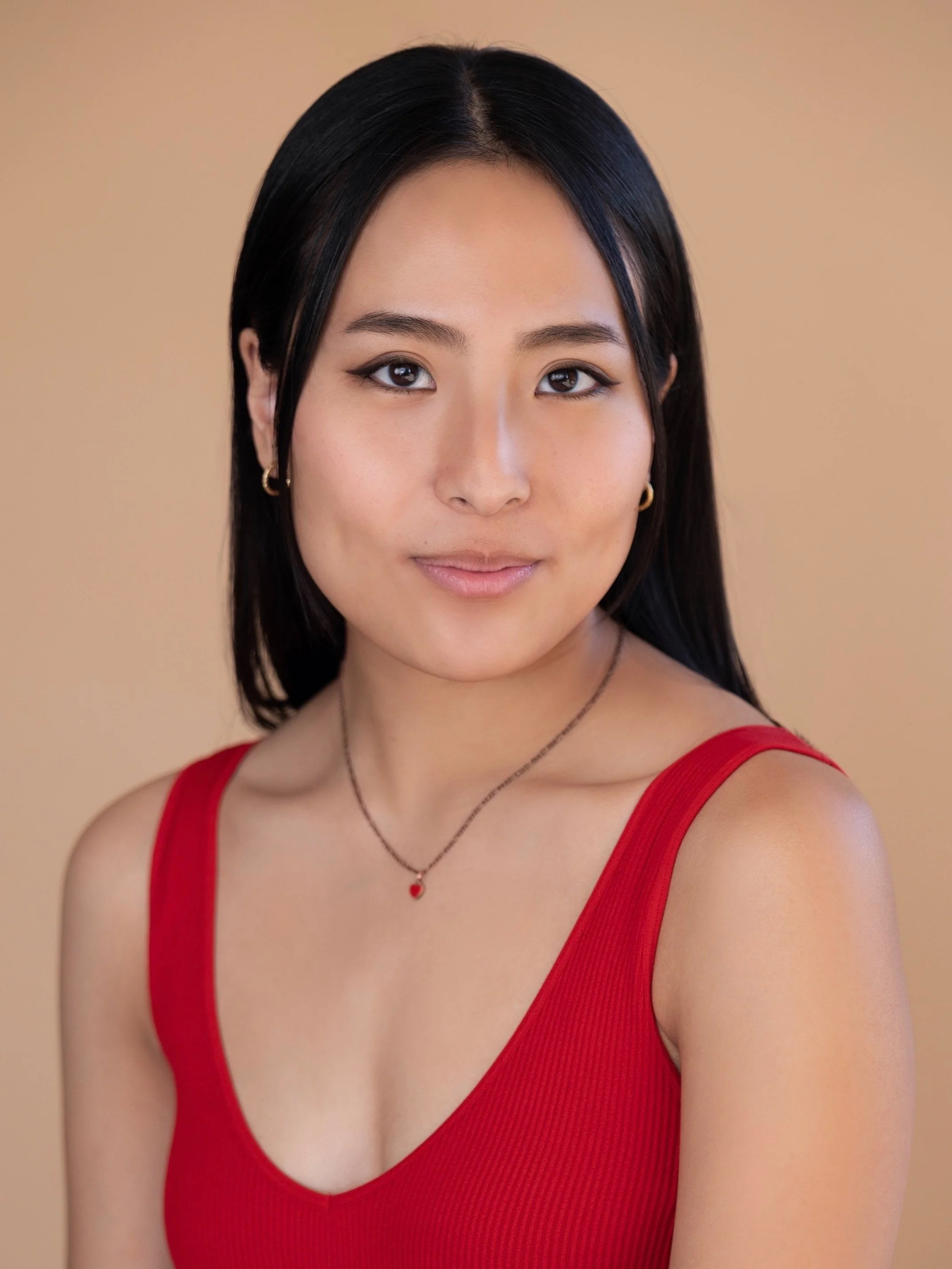 A young woman with straight black hair, wearing a red sleeveless top, gold hoop earrings, a delicate necklace with a red heart pendant, and subtle makeup, standing against a beige background.
