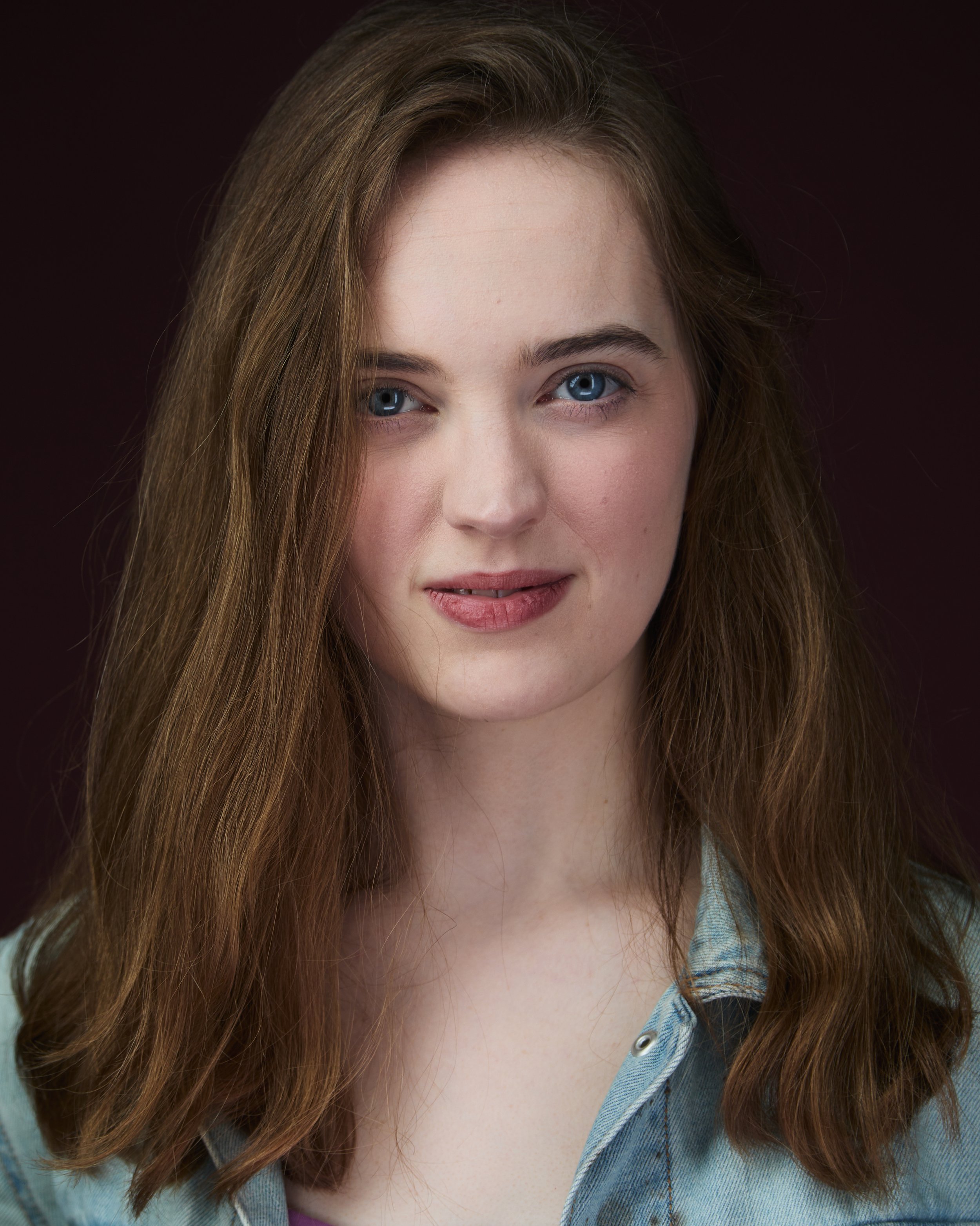 A portrait of a young woman with long light brown hair, blue eyes, wearing a denim shirt, against a dark background.