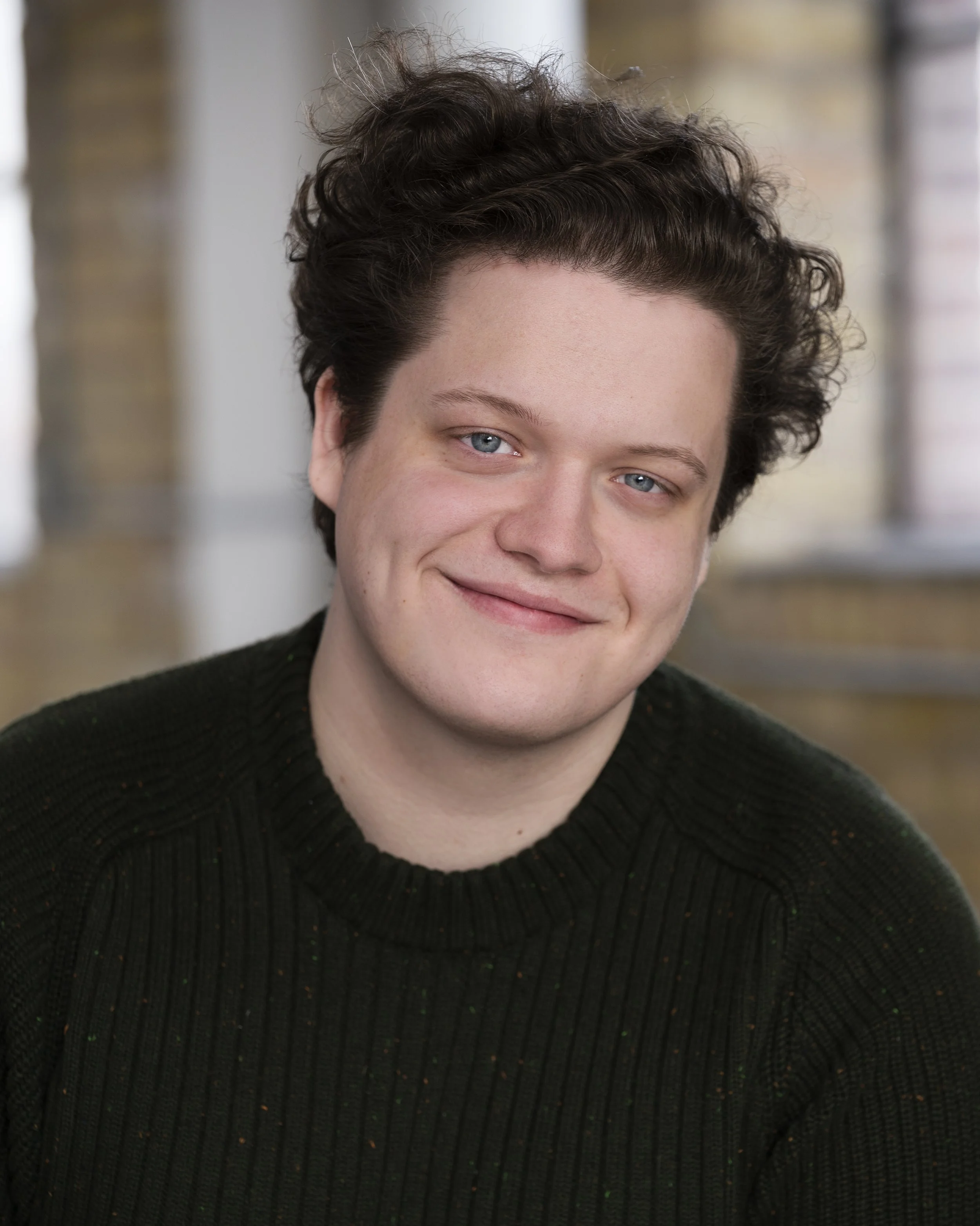A young man with curly brown hair, blue eyes, and fair skin smiling, wearing a black sweater, in an indoor setting with large windows and a brick wall in the background.