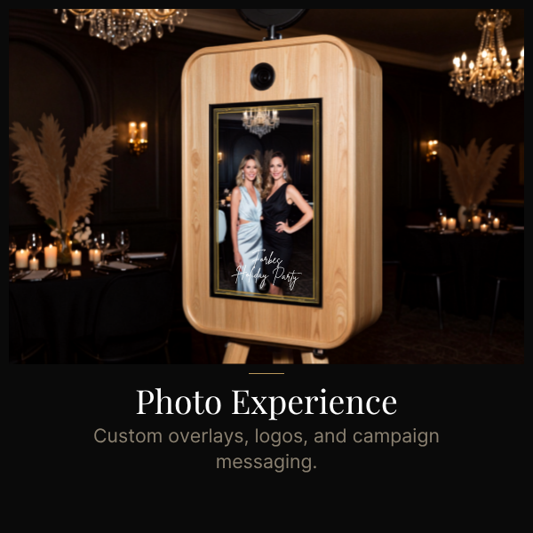 A photo booth with a wooden frame showing a picture of two women at a party, set in an elegant venue with dim lighting, chandeliers, and decorated tables.