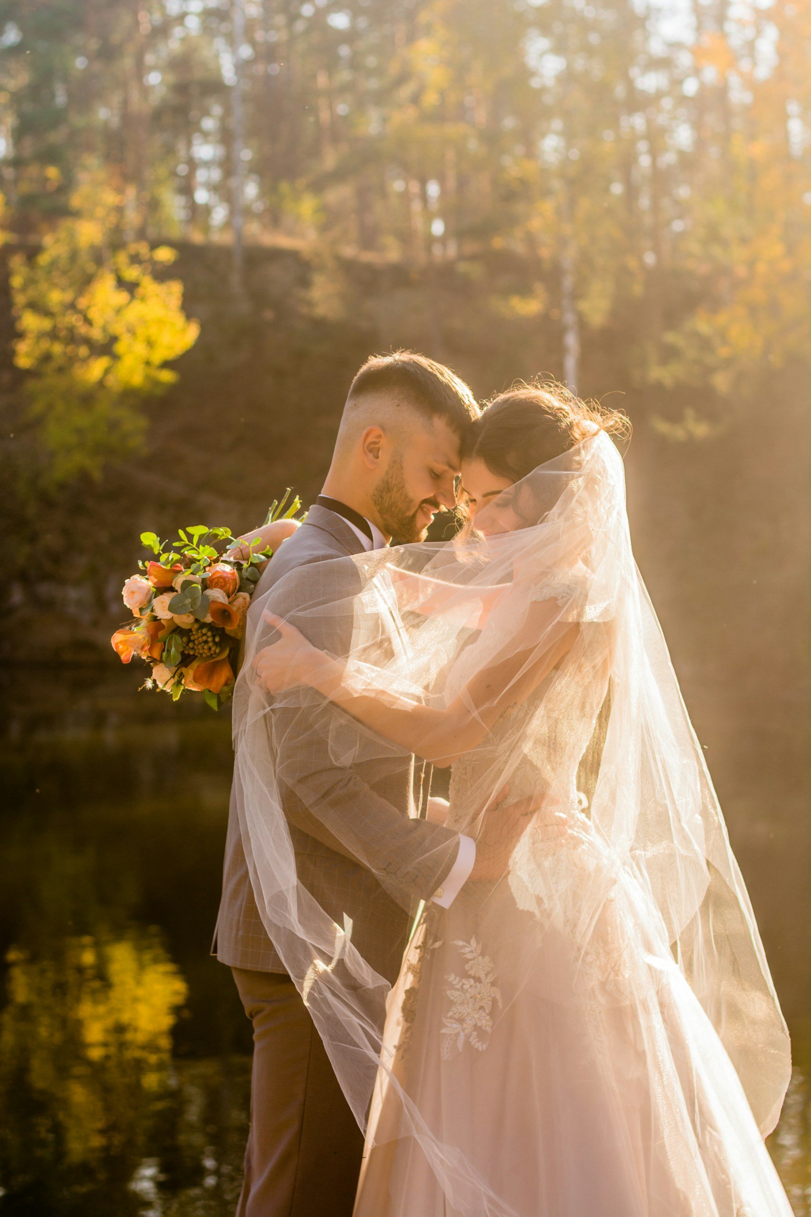 A newlywed couple embracing near a body of water during sunset, with trees in the background, and the bride holding a bouquet of flowers.