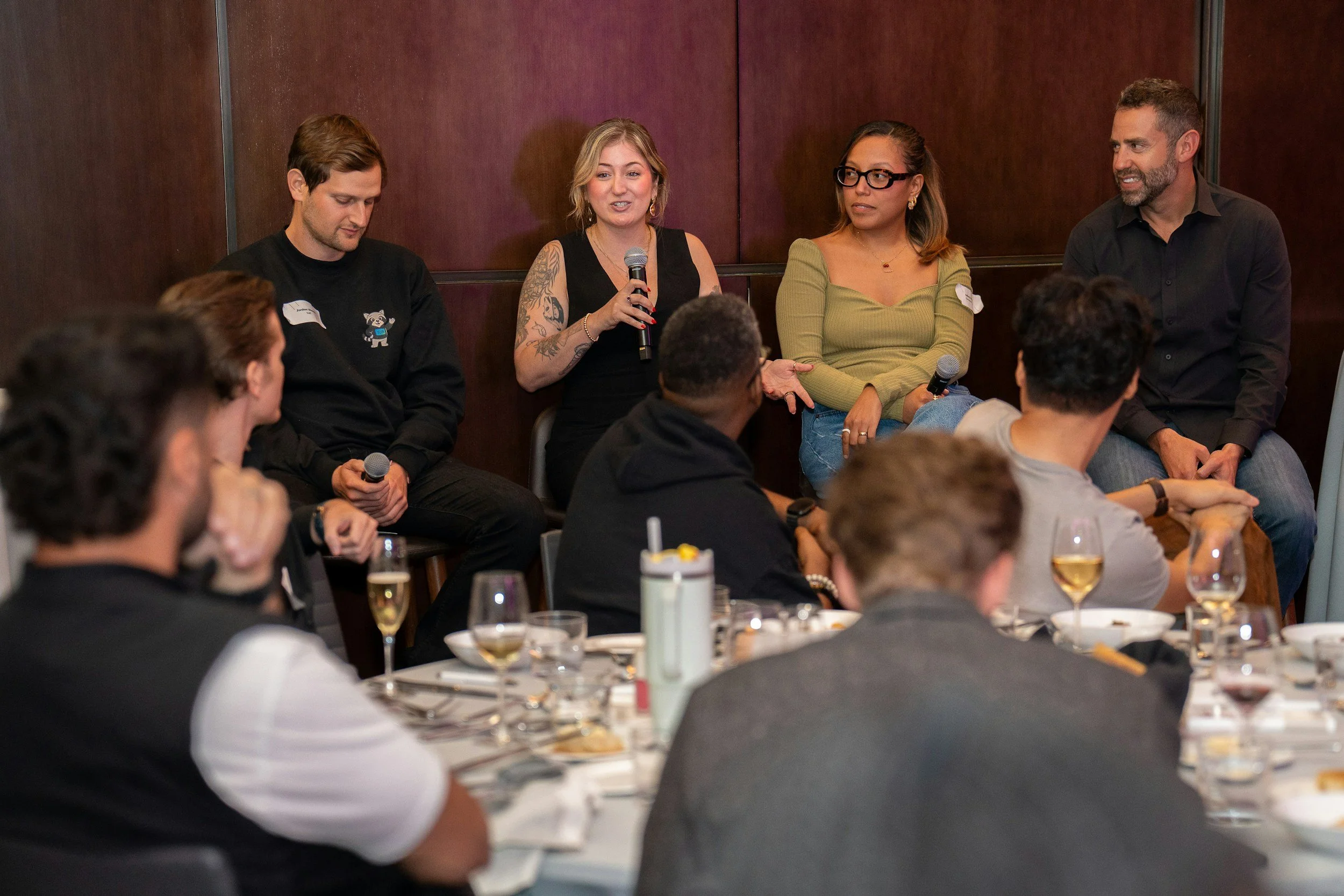 Four individuals are seated in a panel discussion at an event. The woman second from the left is speaking into a microphone, with others listening. The audience is visible in the foreground, and there are tables with drinks and food.