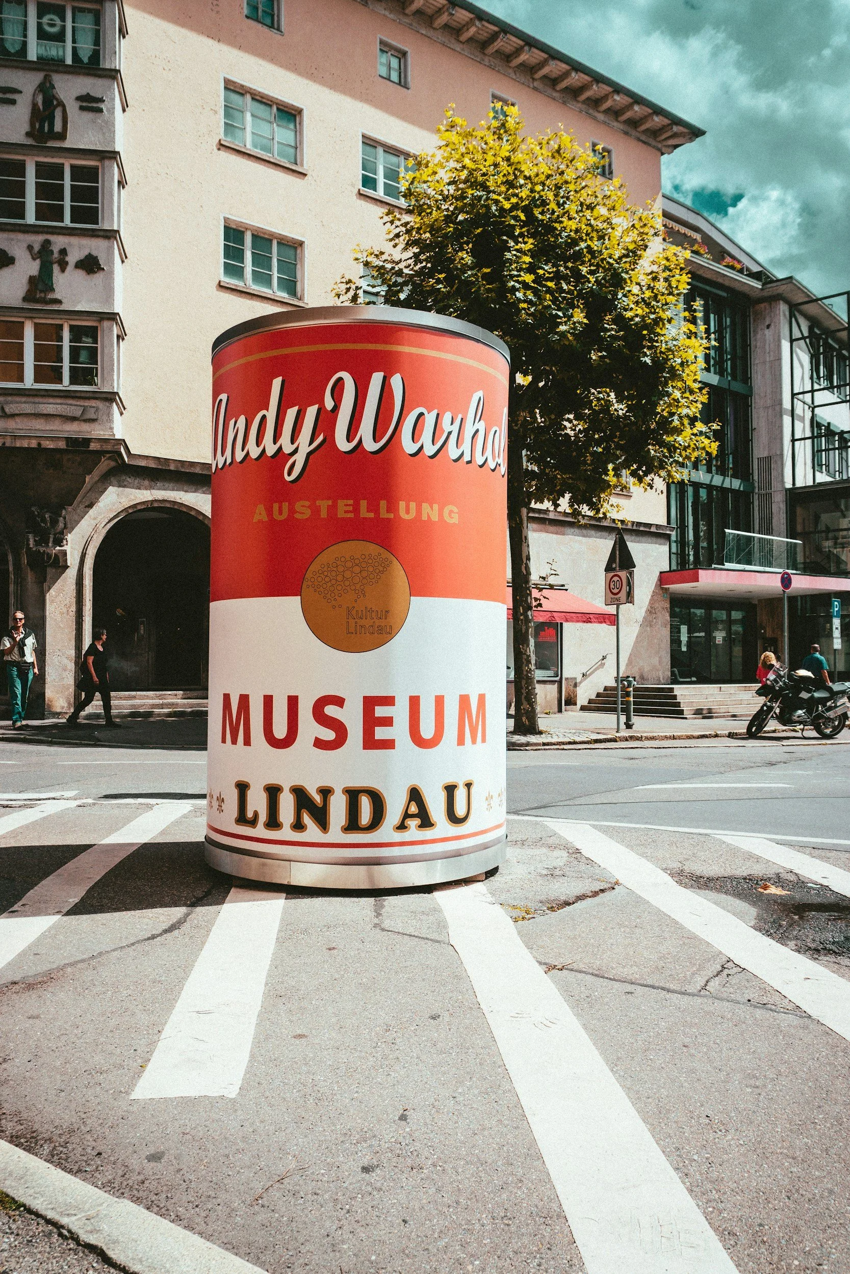 A large cylindrical signboard on a city street advertising an exhibition at the Kultur Lindau Museum, with a historic building, people walking and a motorcycle in the background.