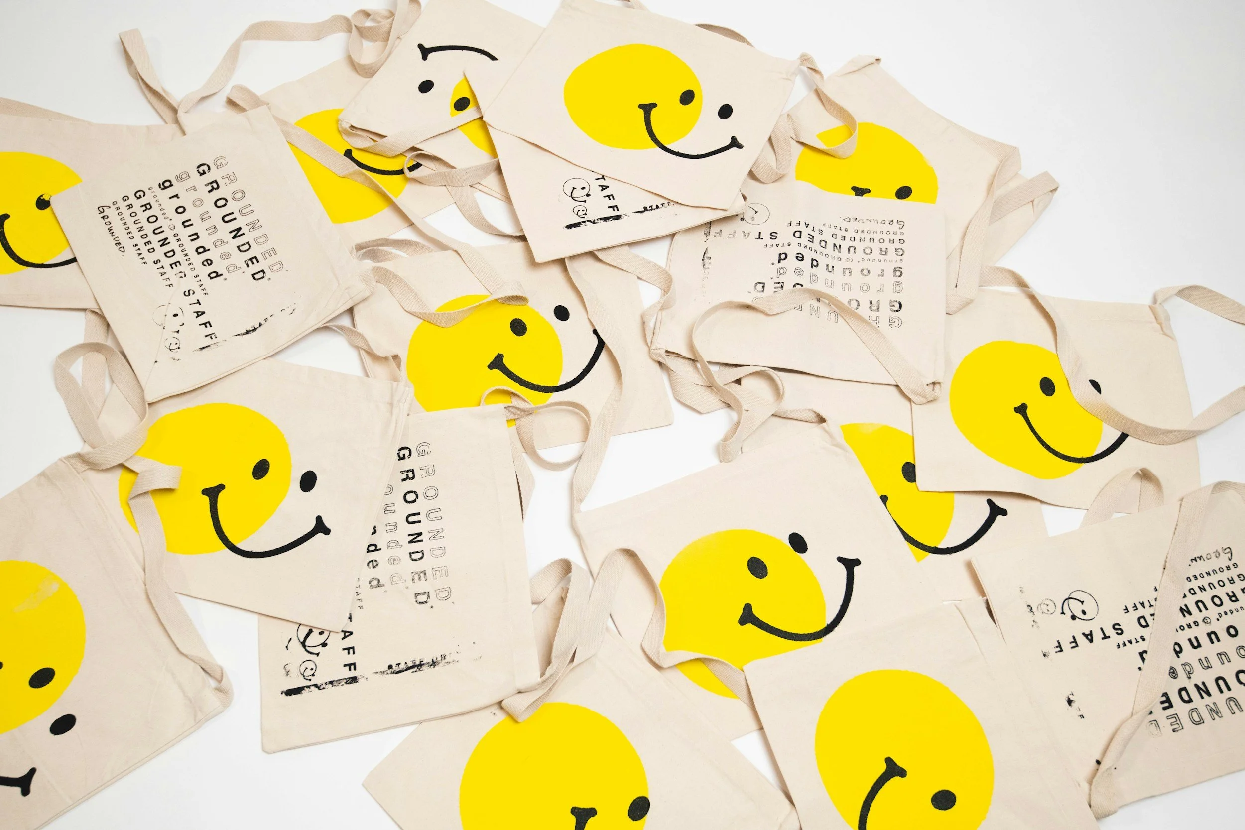 Multiple reusable cloth tote bags with a yellow smiley face Design and printed text, scattered on a white surface.