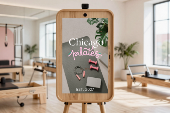A digital sign in a Pilates studio displays the text 'Chicago Pilates' with a pink Pilates logo and the year 'EST. 2027', with Pilates equipment and plants in the background.