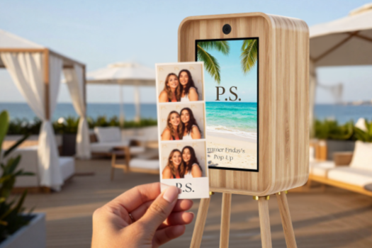 A person holding two photo booth pictures in front of a wooden photo kiosk with a beach scene on the screen. The background features a seaside outdoor lounge with white canopies and umbrellas.