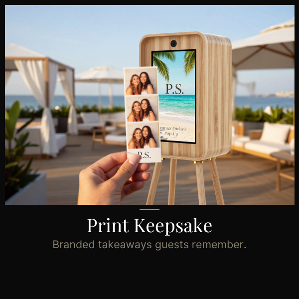 A person holds a photo booth print strip in front of a digital photo booth displaying a beach scene with palm trees and the initials 'P.S.' at a tropical outdoor venue with white canopies and lounge furniture.
