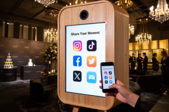 A digital social media sharing station resembling a large wooden photo booth, displaying icons for Instagram, TikTok, Facebook, Twitter, X, and email, with a person holding a smartphone to share content, in an elegant event space with chandeliers and a cake table.