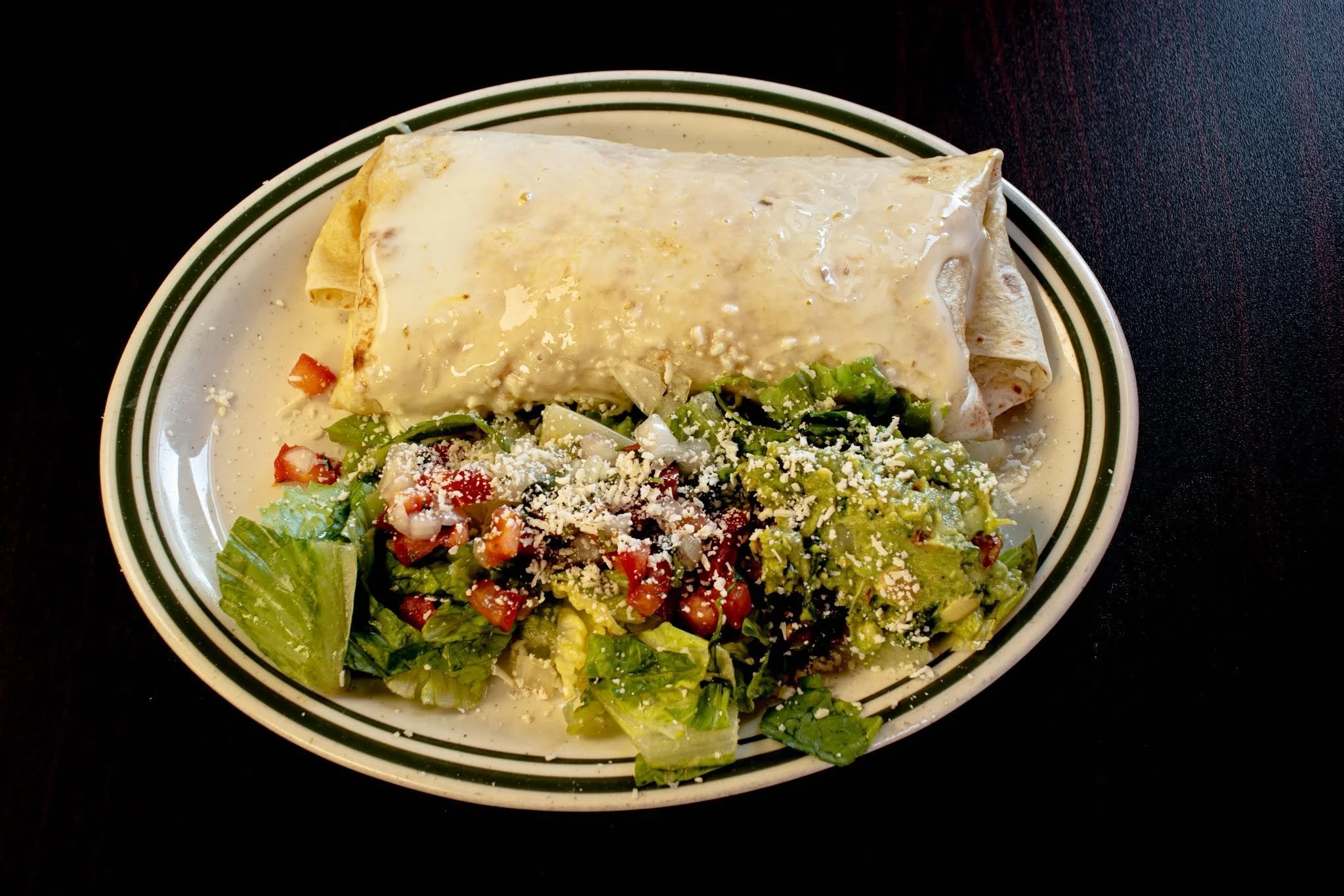 A plate with a folded burrito covered in white sauce, accompanied by a side of chopped lettuce, tomatoes, guacamole, and shredded cheese.
