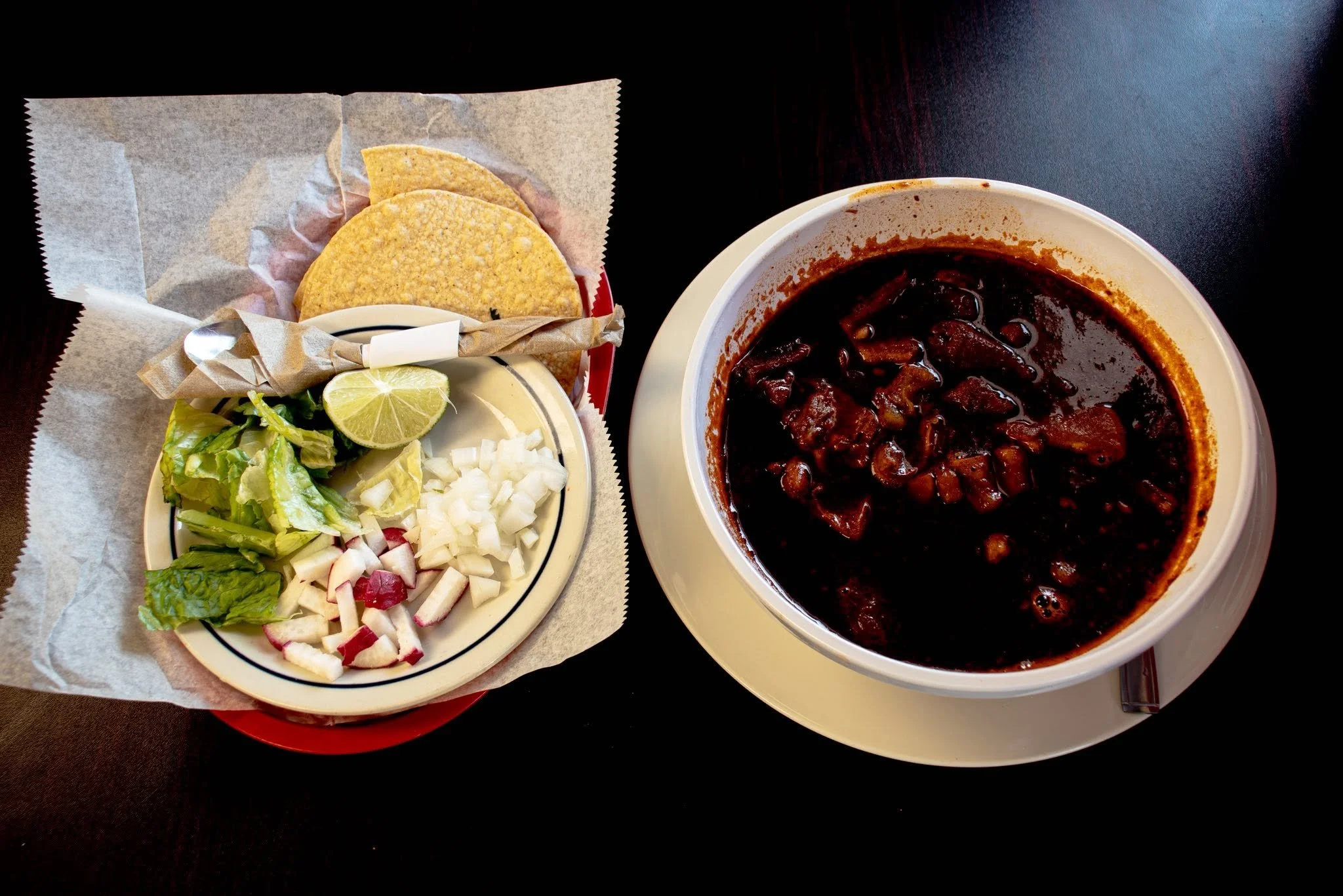 A plate of salad with lettuce, radishes, onions, lemon wedges, and two taco shells, alongside a bowl of dark, rich chili with chunks of meat and beans.