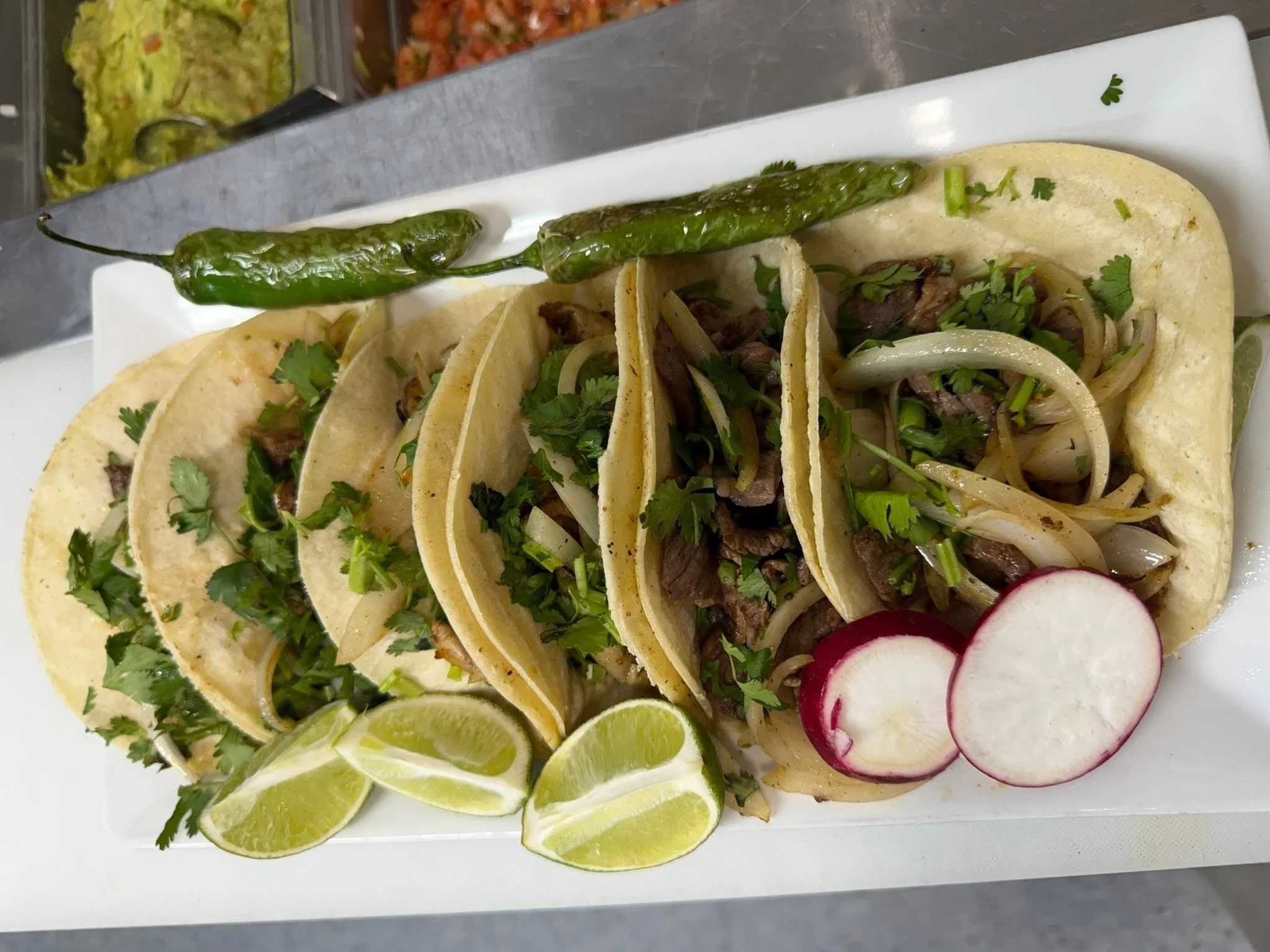 Close-up of three tacos filled with meat, onions, cilantro, and sliced radishes on a white plate, garnished with lime wedges and grilled green chilies, with traditional Mexican toppings in the background.