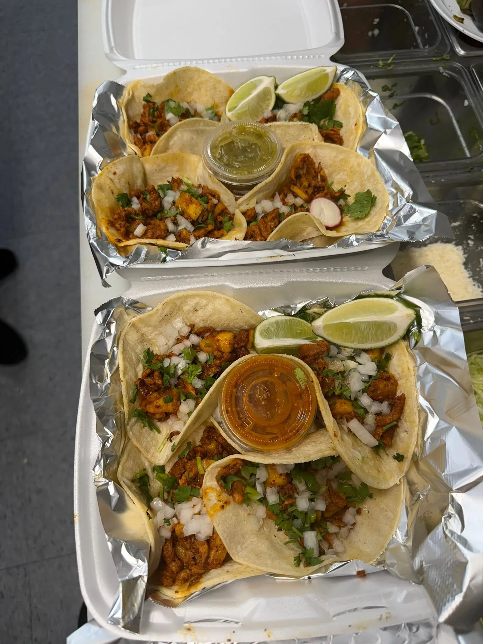 Four tacos with seasoned meat, chopped onions, cilantro, and lime wedges served on a foil-lined tray, with small containers of salsa and dipping sauce.