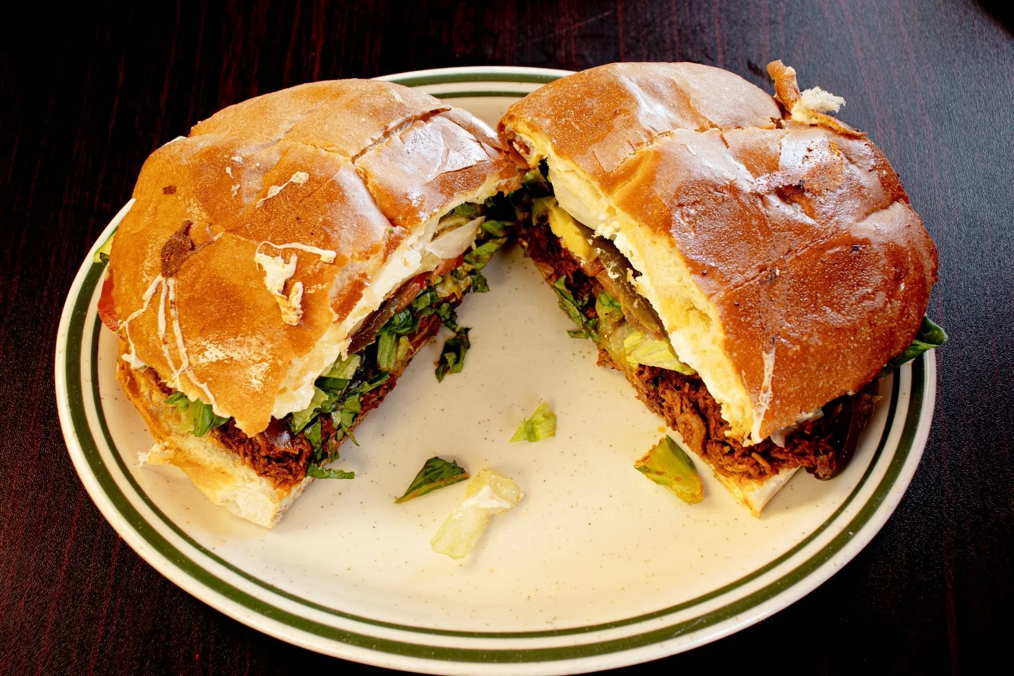 A sandwich on a plate with a side of green lettuce, sliced in half to show meat, cheese, lettuce, tomato, and bread, with some lettuce and bread crumbs around.