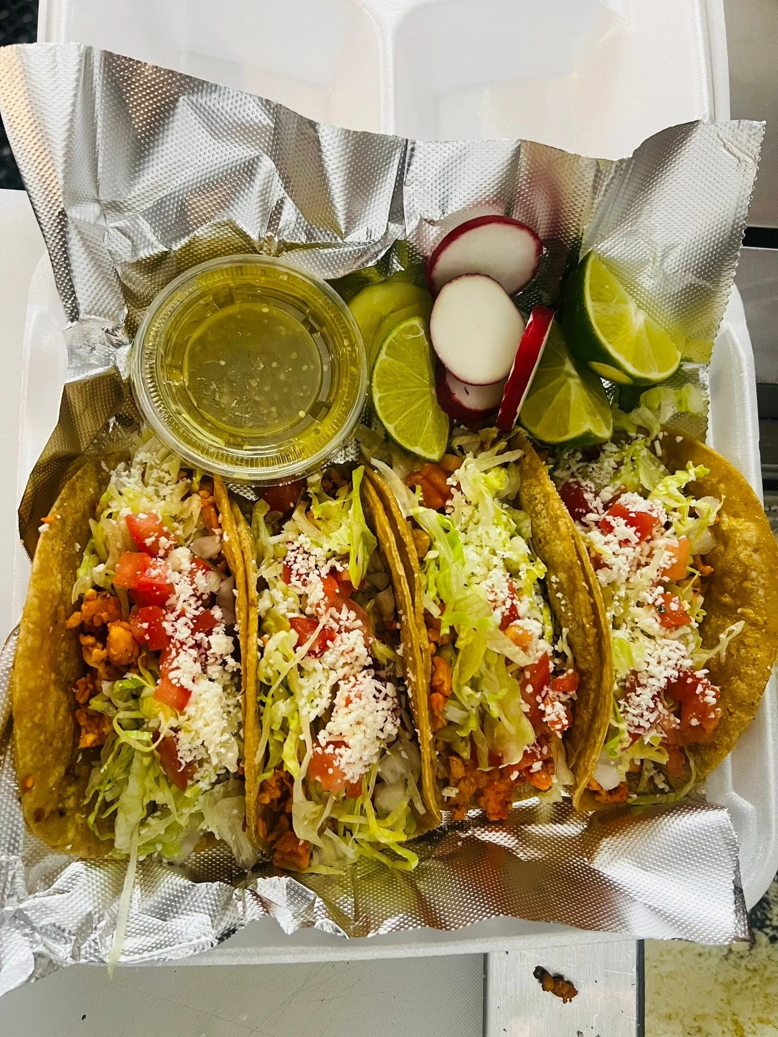 Three tacos filled with chopped lettuce, diced tomatoes, and shredded cheese, placed on a foil-lined tray with lime wedges, radish slices, and a small cup of salsa or sauce.