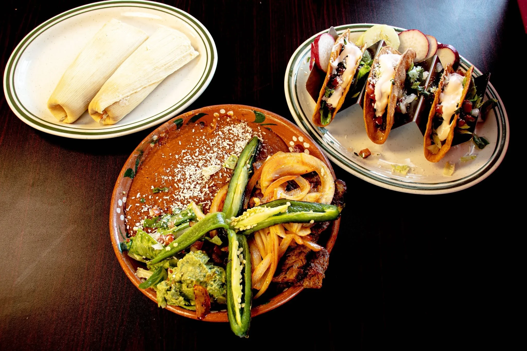 Plate of three Mexican tacos with meat, lettuce, cheese, and sauce, and a bowl of refried beans topped with cheese, on a dark table.