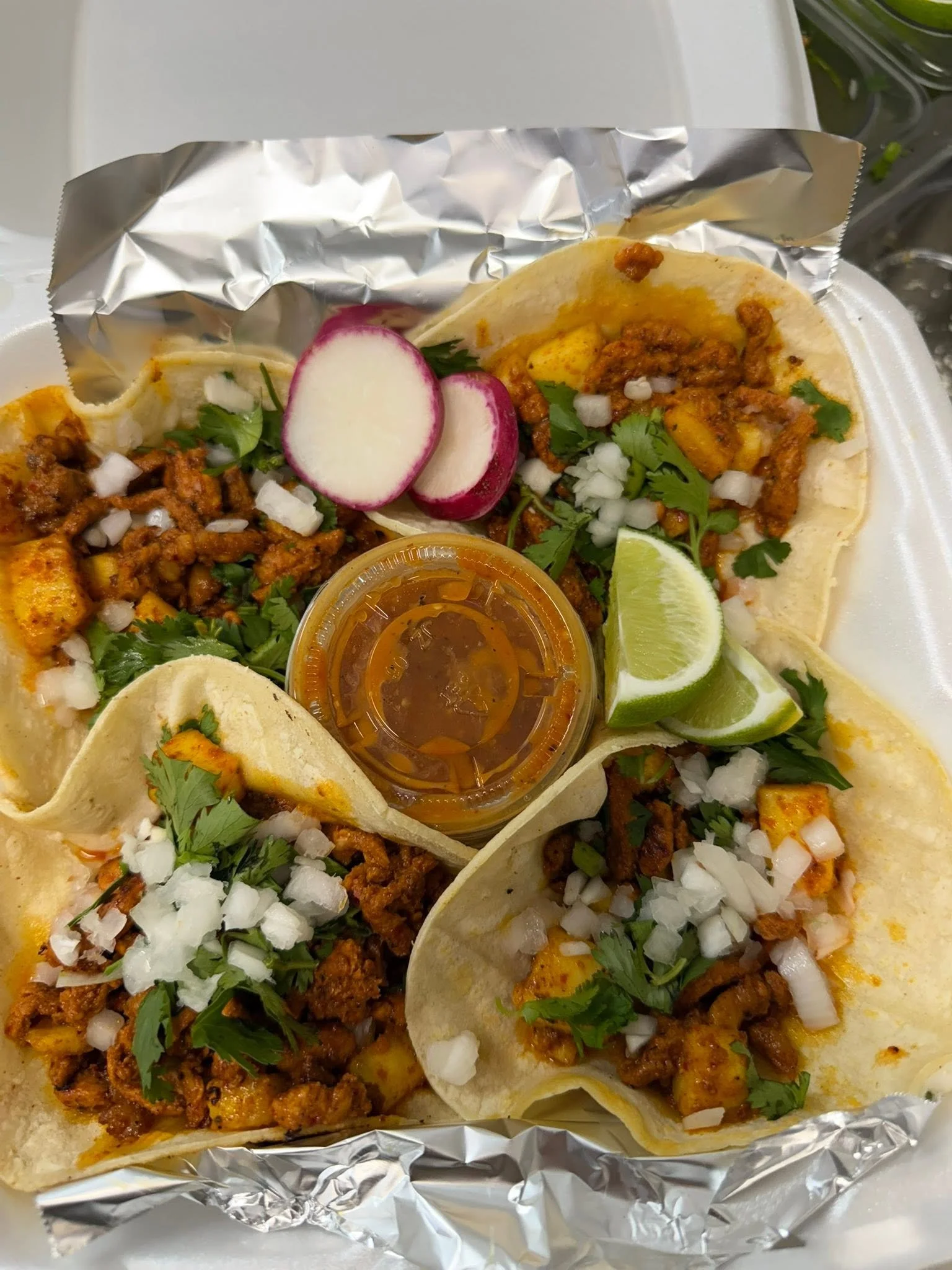 Close-up of three beef tacos with chopped onions, cilantro, radishes, lime wedges, and a small container of salsa on foil-lined tray.