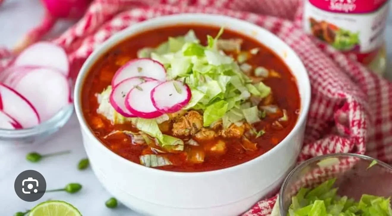 A bowl of spicy red soup topped with shredded lettuce and sliced radishes, with radishes and lime slices on the side.